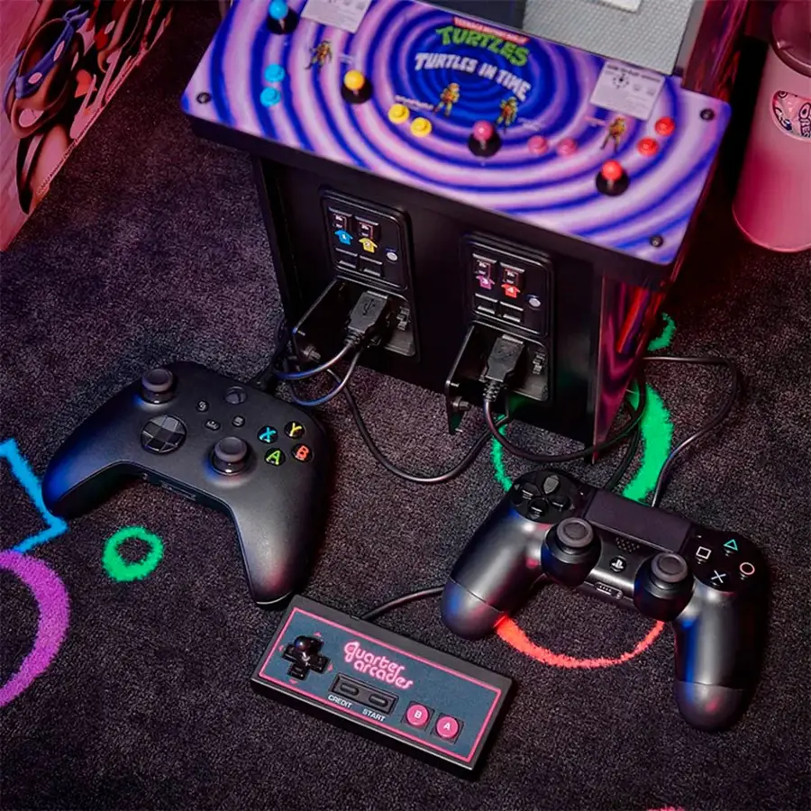 The image features a Turtles in Time arcade machine with a purple and black color scheme. There are two game controllers placed on the floor in front of the machine, and a couple of additional controllers are also visible in the scene. The machine is connected to a TV, which is located on the left side of the image. The overall setting seems to be a casual gaming environment, with the controllers and machine ready for use.