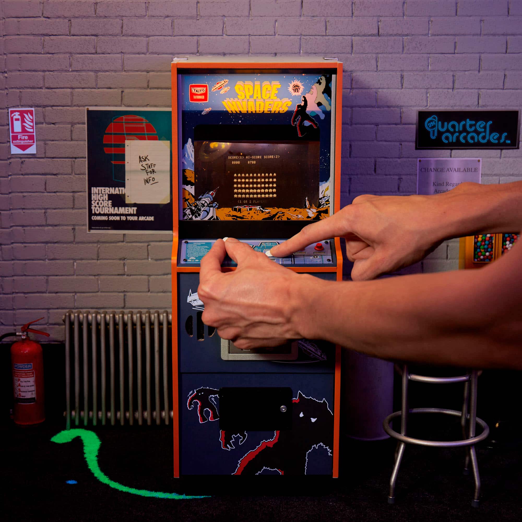 The image features a person playing a video game on a Space Invaders machine. The machine is located in a room with a brick wall and a heater. The person is using a joystick to control the game, and there is a fire extinguisher nearby. The room also has a chair and a dartboard. The Space Invaders machine is an old-fashioned arcade game, and the person is likely enjoying a nostalgic gaming experience.
