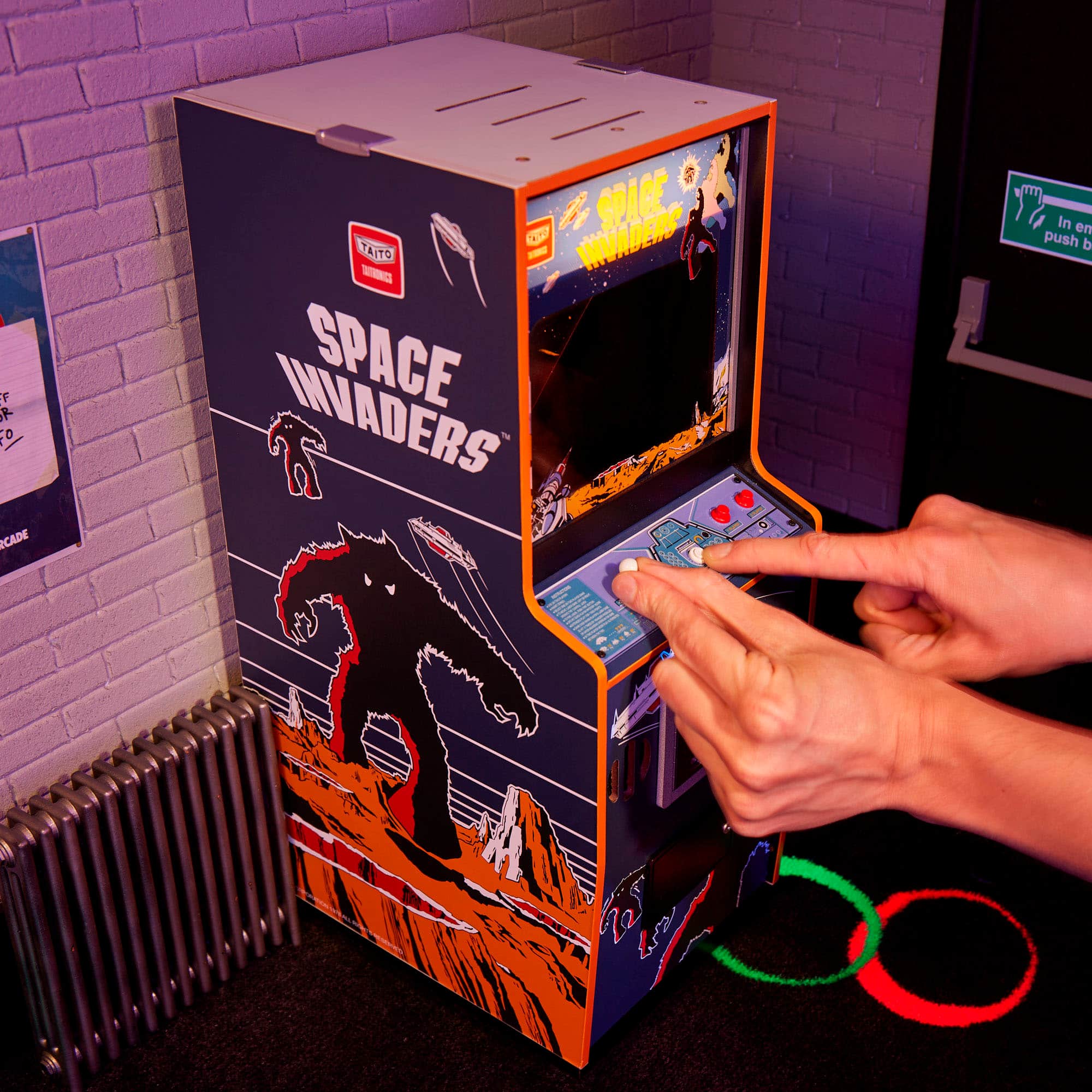 The image features a person playing a video game, specifically Space Invaders, on a small arcade machine. The machine is located on a table, and the person is using a joystick to control the game. The game is displayed on a screen, and the person's hand is visible, pointing at the screen. The arcade machine is designed to be compact and accessible for players of all ages.