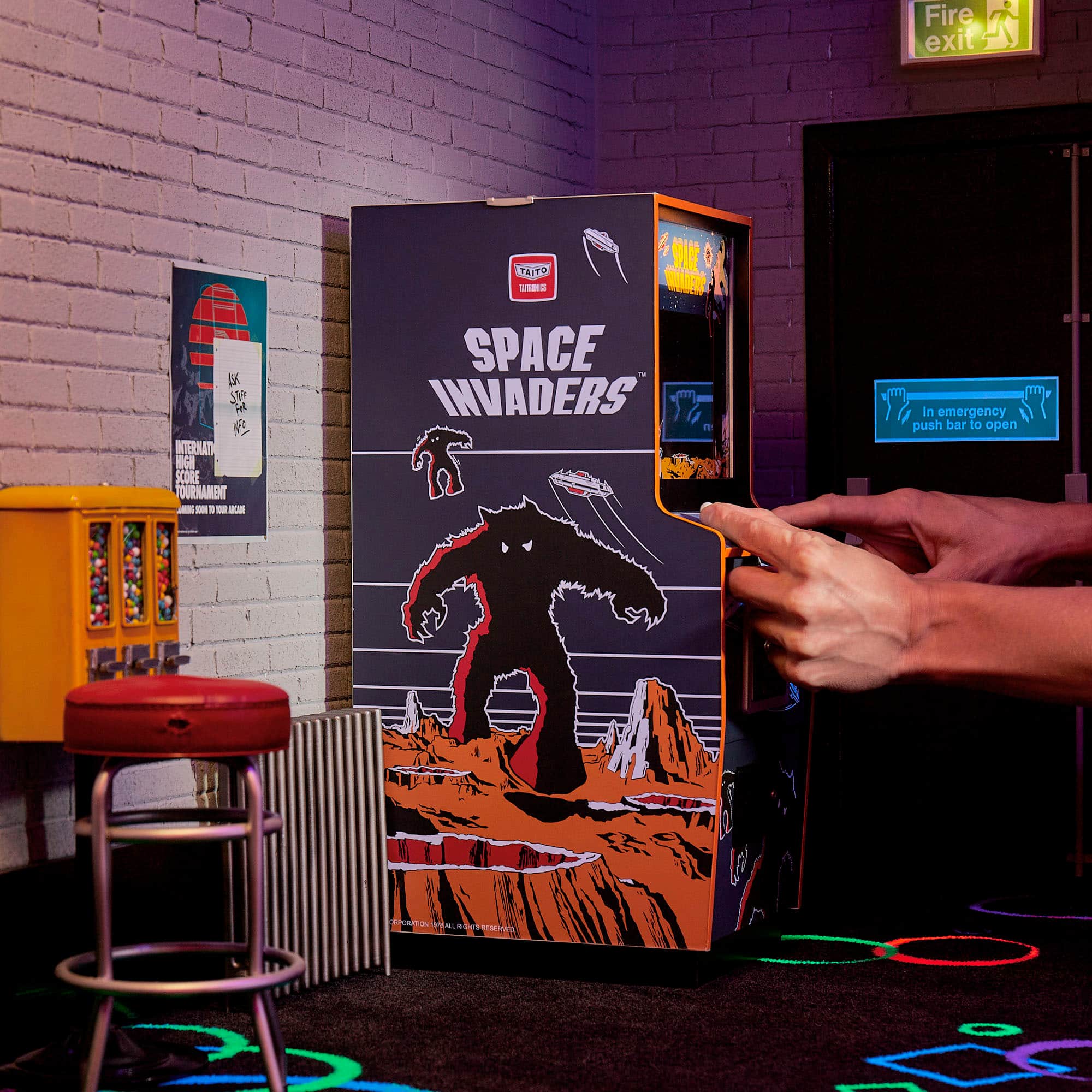 The image features a Space Invaders arcade machine with a large monster on the front. A person is playing the game, pressing a button on the machine. The arcade machine is located in a room with a brick wall and a fire exit sign nearby. The room also has a chair and a bench, providing seating options for players.