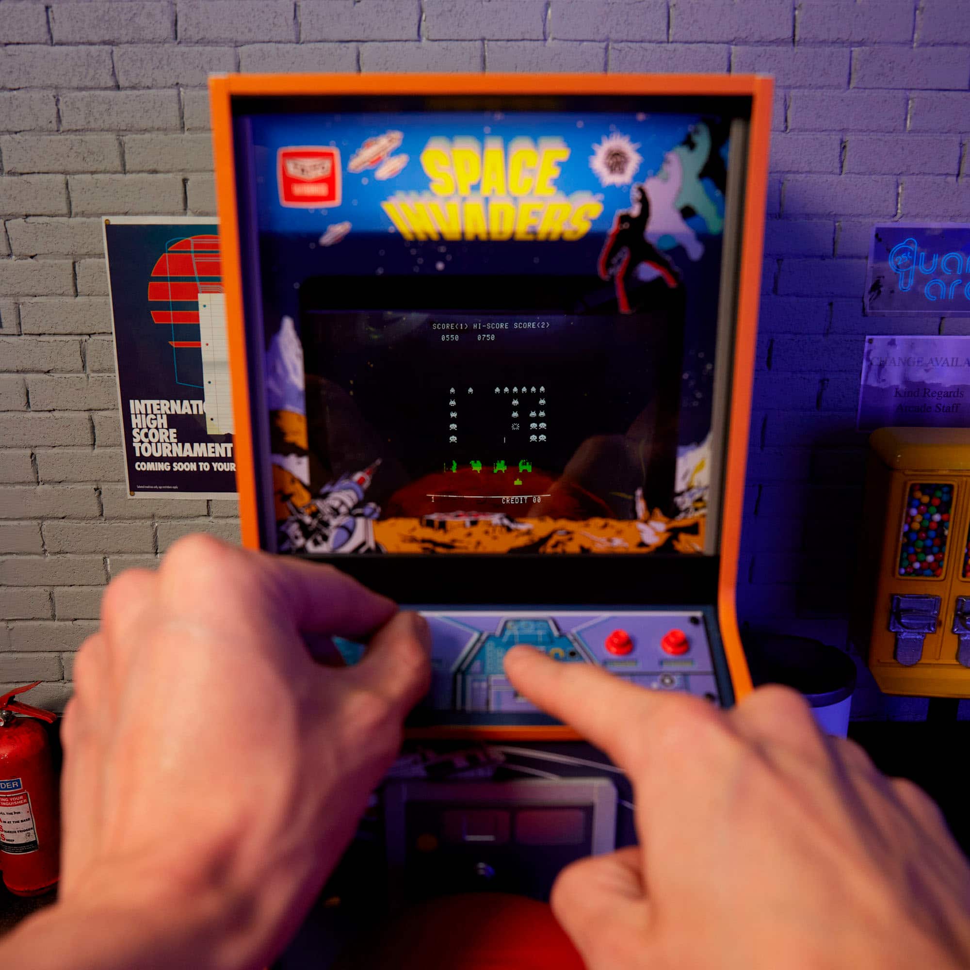 The image features a person playing a video game, specifically Space Invaders, on an old-fashioned arcade machine. The game is displayed on a screen, and the person is using a joystick to control the game. The arcade machine is placed in a brick-walled room, and there is a fire extinguisher nearby. The image also includes a poster on the wall, possibly advertising the game or the arcade itself.