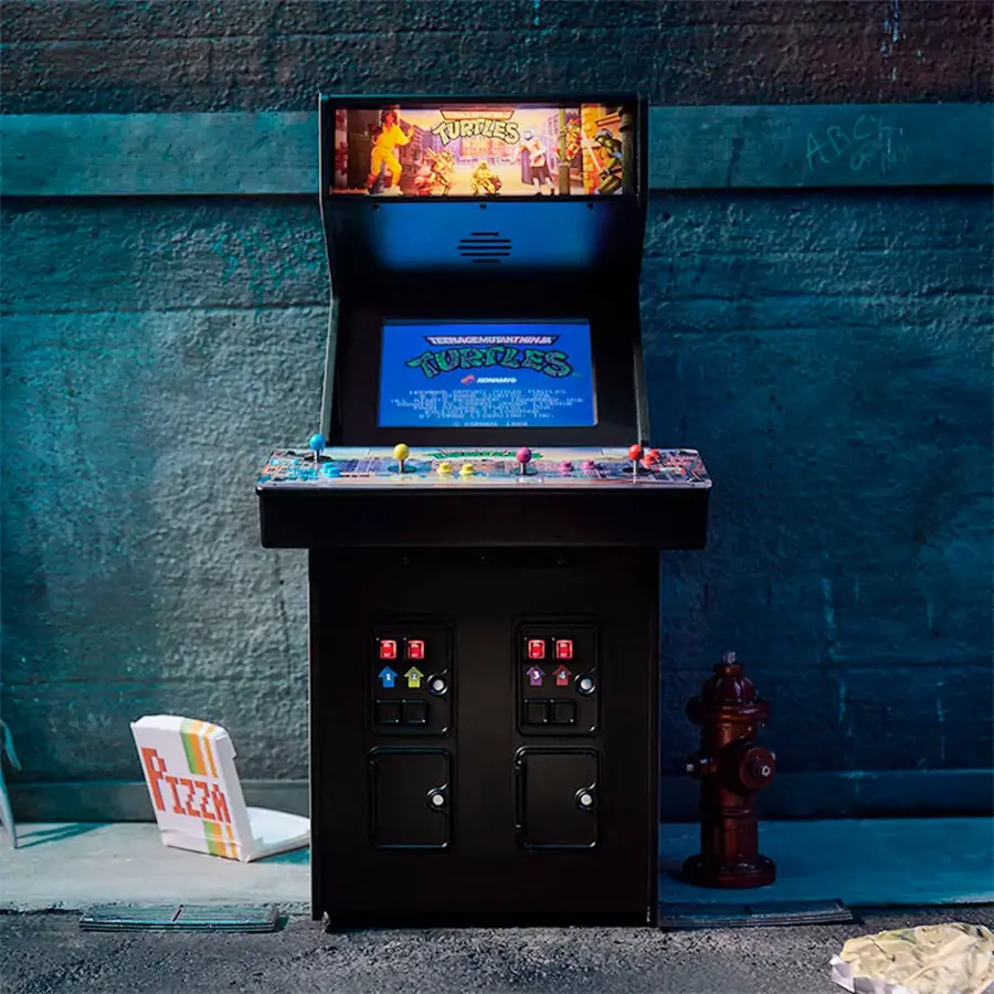 The image features a black arcade machine, likely a Teenage Mutant Ninja Turtles game, sitting on the sidewalk next to a fire hydrant. The machine is positioned in front of a building, and there is a pizza box nearby.