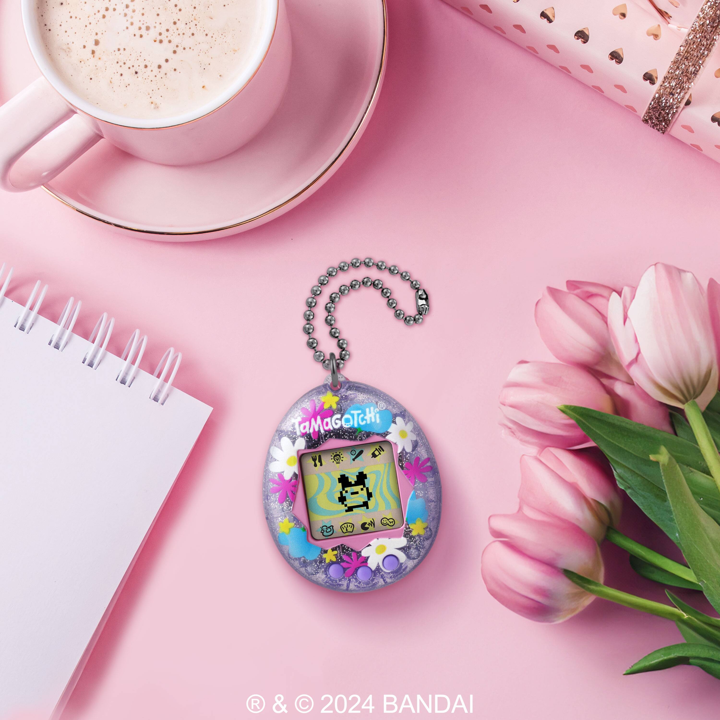 The image features a pink background with a notebook, a coffee cup, and a small cat charm. The charm is hanging from a chain and is placed near the coffee cup. The scene also includes a vase with pink flowers, adding a touch of color and elegance to the setting.