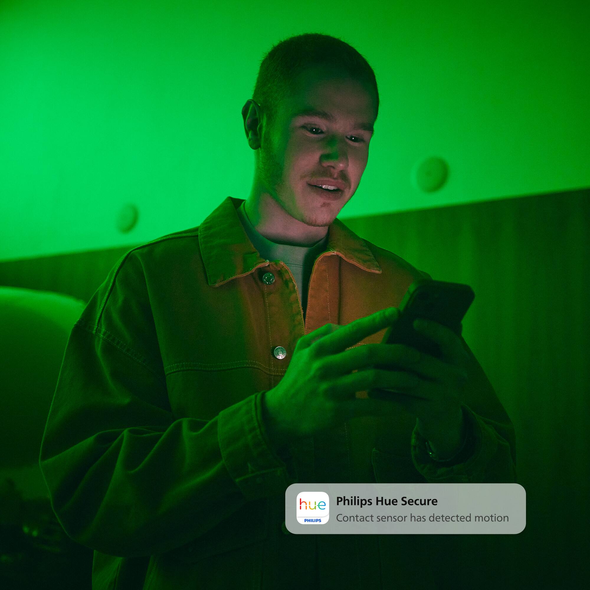 The image shows a man wearing a yellow jacket and holding a cell phone in his hand. The man appears to be looking at his phone, possibly checking messages or browsing the internet. The scene is set in a dimly lit room, with the man standing in the center of the frame. The image also features a Philips Hue Secure sensor, which is a motion-detecting device that helps ensure the security of the home.