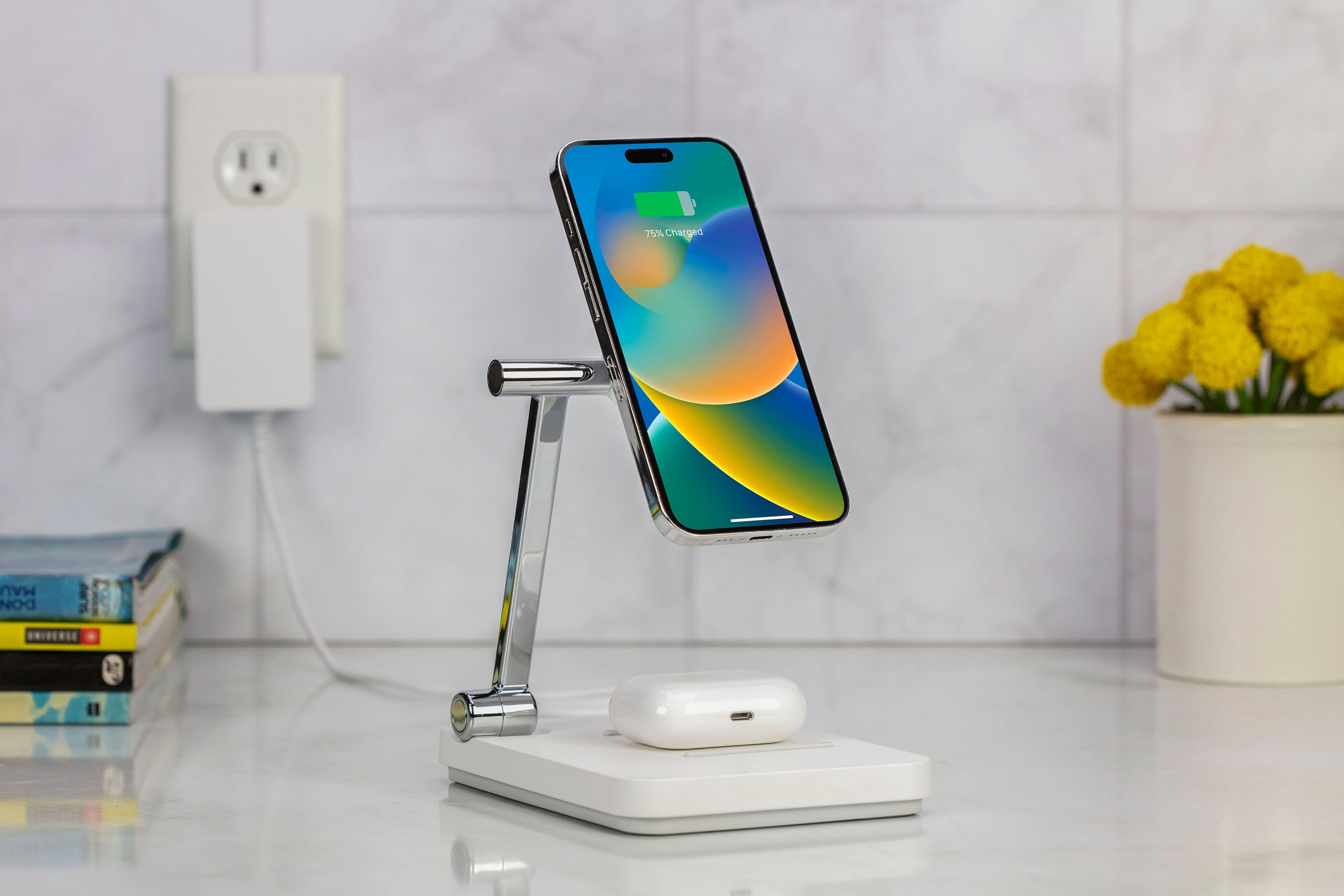 The image shows a smartphone on a stand, which is placed on a table. The phone is 75% charged, and it is a model of the iPhone. The stand is designed to keep the phone upright and easily accessible. The table is white, and there are some books nearby. Additionally, there are yellow flowers in a vase, adding a touch of color and life to the scene.