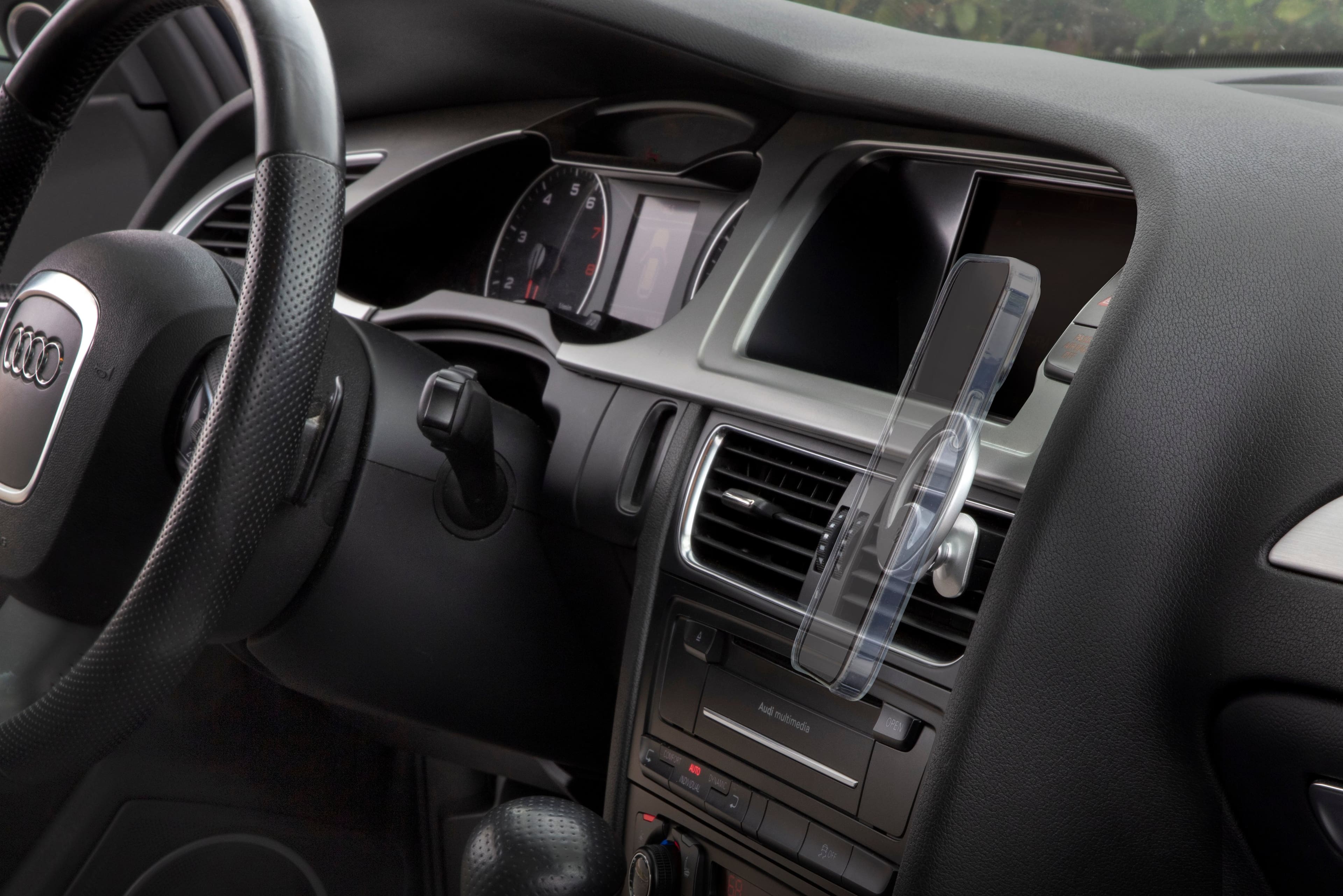 The image shows the interior of a car with a steering wheel and a dashboard. The dashboard has a clock and a speedometer, and there is a small car air freshener hanging from the dashboard. The car is equipped with a radio, and the steering wheel is positioned close to the dashboard. The car appears to be a luxury model, possibly an Audi, with a focus on the multimedia system.