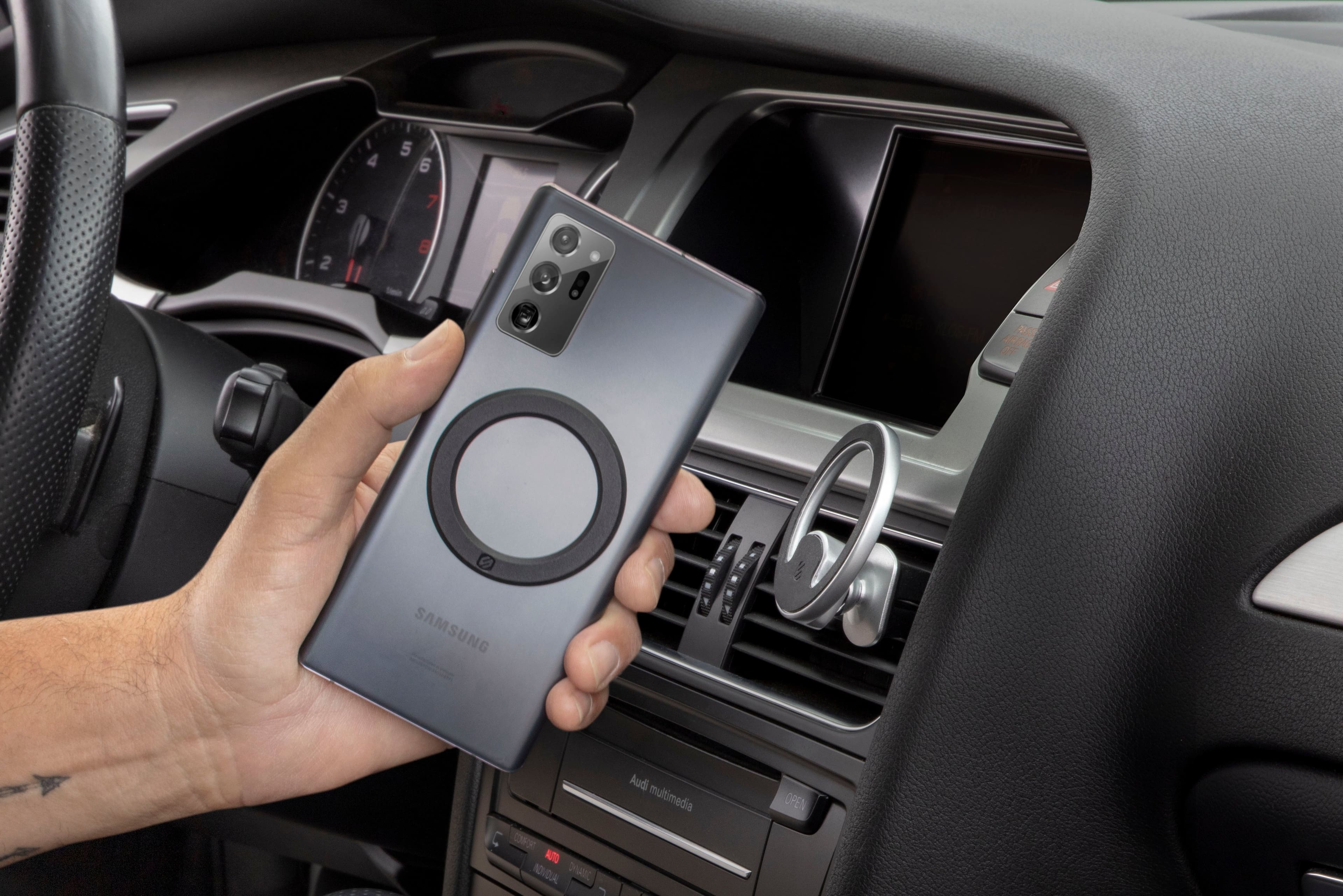 The image shows a person holding a Samsung phone in their hand while sitting in a car. The car has a dashboard with a clock and a steering wheel. The person is likely using the phone to control the car's entertainment system or to access information related to the car's features. The car appears to be a modern model with advanced technology and multimedia capabilities.