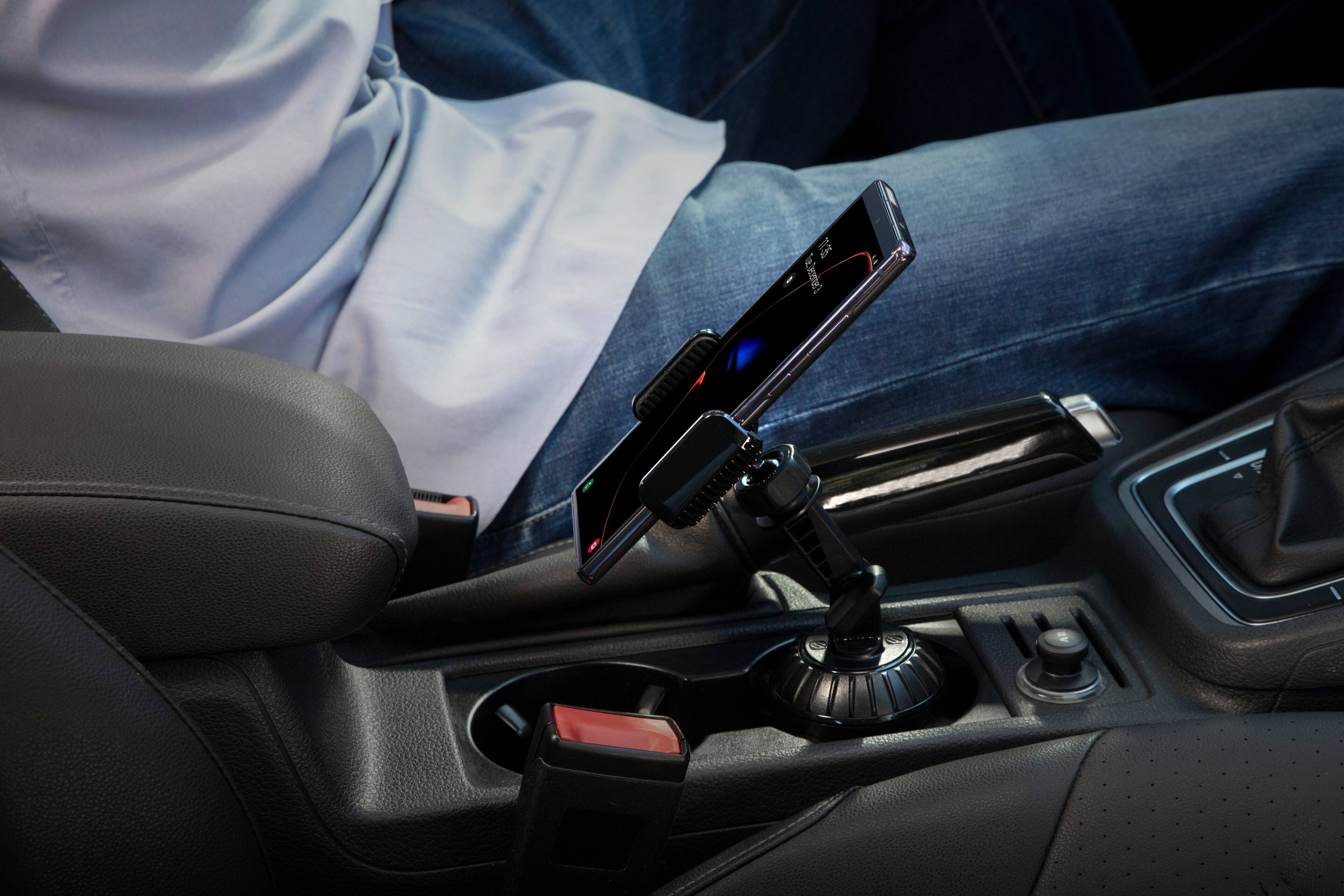 The image shows a person sitting in a car with a cell phone mounted on the dashboard. The cell phone is positioned in a holder, which is attached to the car's dashboard. The person is sitting in the driver's seat, and the car appears to be parked. The cell phone is placed in a holder, which is designed to keep the phone secure and easily accessible while driving. This setup allows the driver to use the phone hands-free, ensuring that their attention remains on the road.