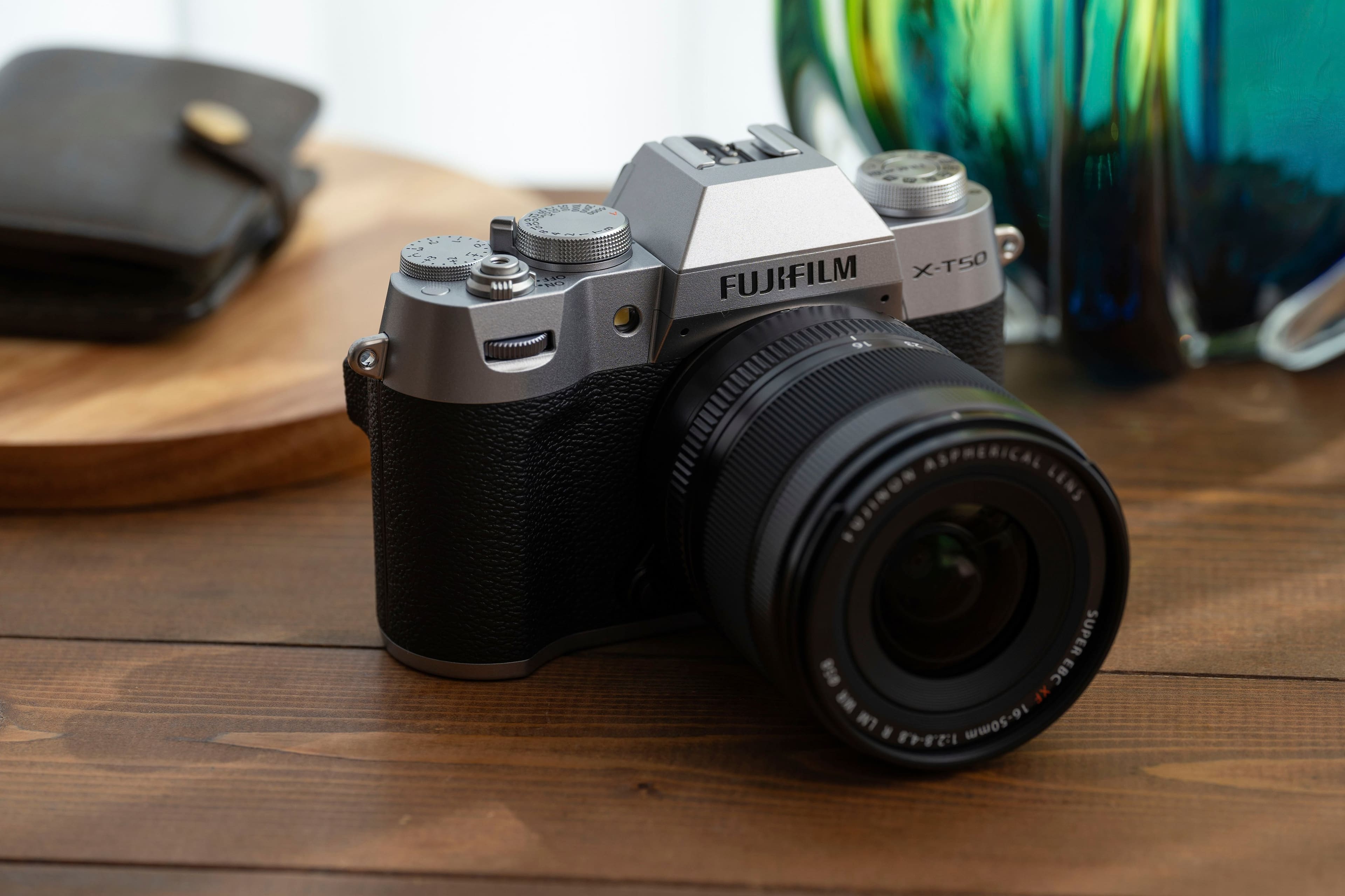 The image features a FUJIFILM X-T50 camera with a black and silver color scheme. The camera is placed on a wooden table, and there is a lens attached to it. The lens is a 16-50mm lens, which is suitable for various photography needs. The camera is positioned on the table, ready to be used for capturing images.