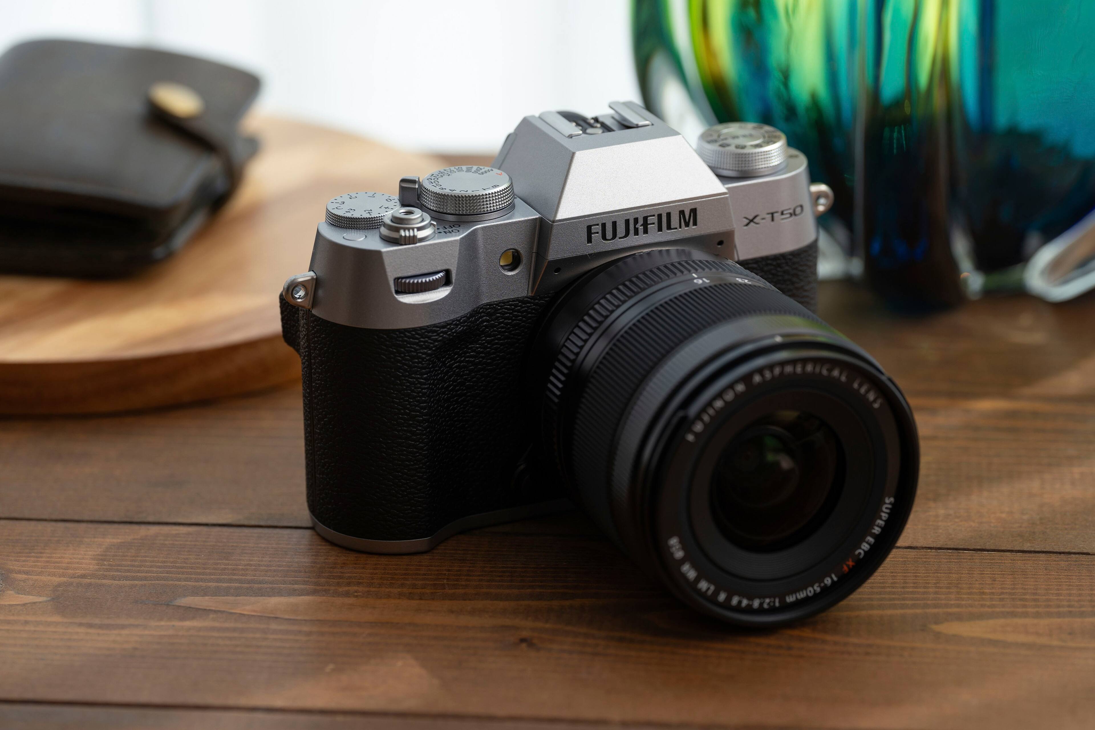 The image features a FUJIFILM X-T50 camera with a black and silver color scheme. The camera is placed on a wooden table, and there is a lens attached to it. The lens is a 16-50mm lens, which is suitable for various photography needs. The camera is positioned on the table, ready to be used for capturing images.