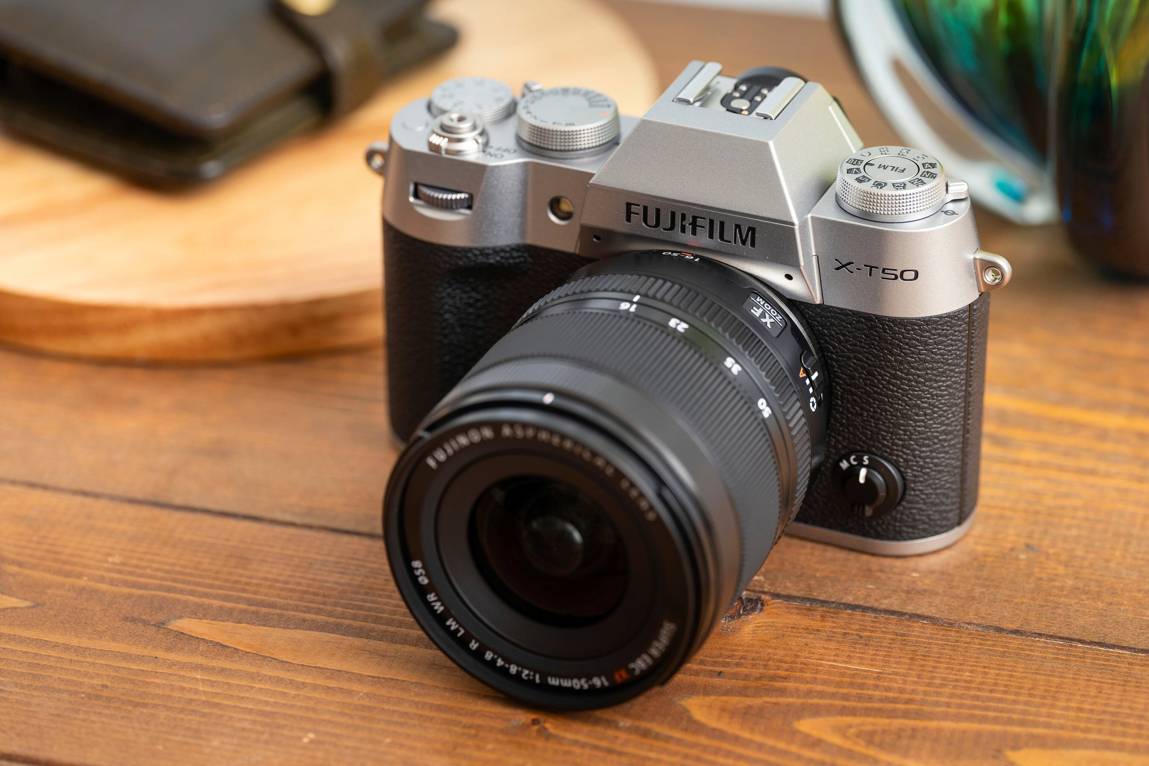 The image features a Fujifilm camera with a large lens sitting on a wooden table. The camera is positioned in a way that the lens is prominently visible, showcasing its size and quality. The camera is likely a high-end model, given its brand and the presence of a large lens.