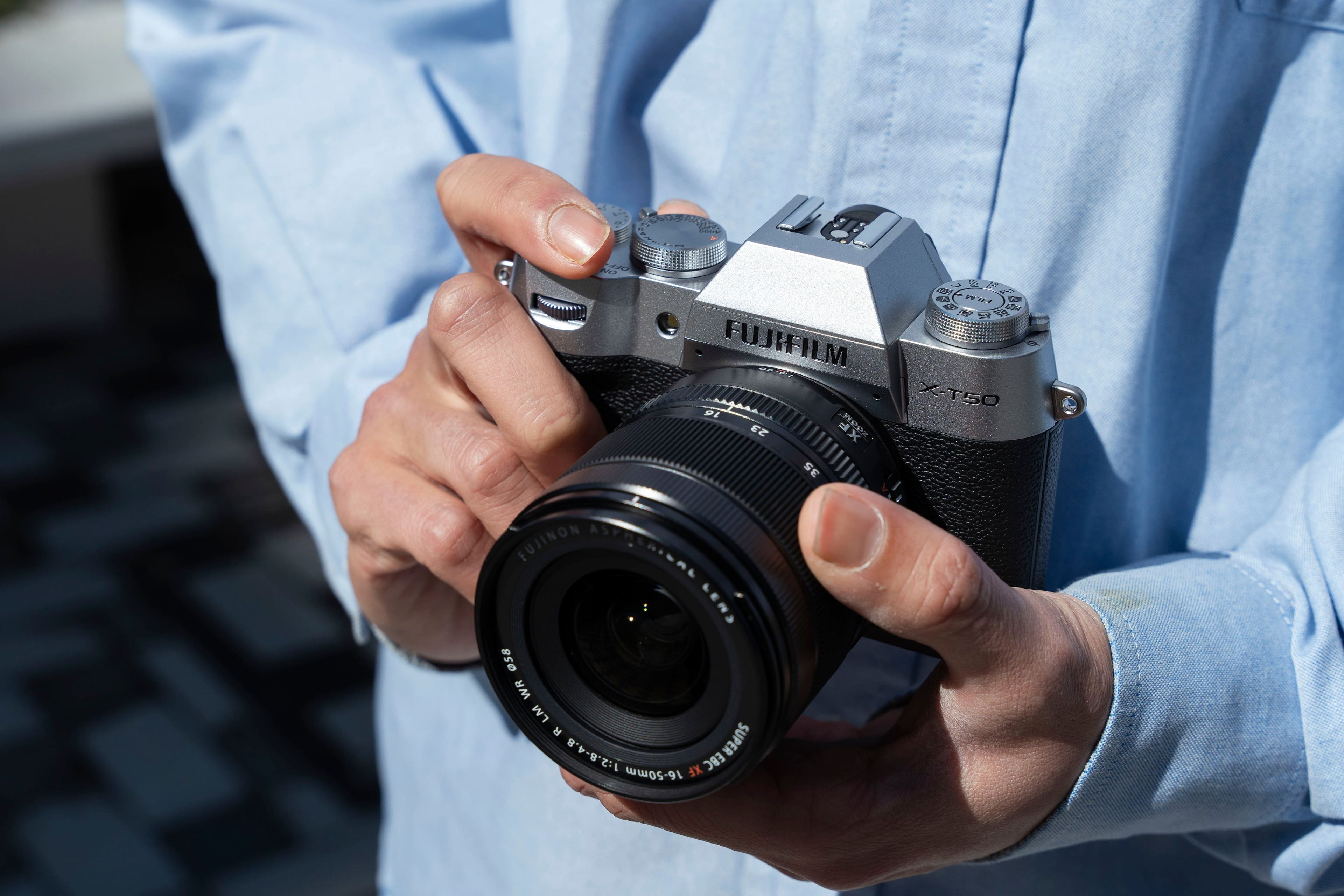 The image features a person holding a Fujifilm X-T50 camera in their hands. The camera is a digital camera with a lens attached to it. The person is likely preparing to take a photograph or has just finished taking one. The camera is a professional-grade device, as indicated by the Fujinon lens and the XF Fujifilm branding.