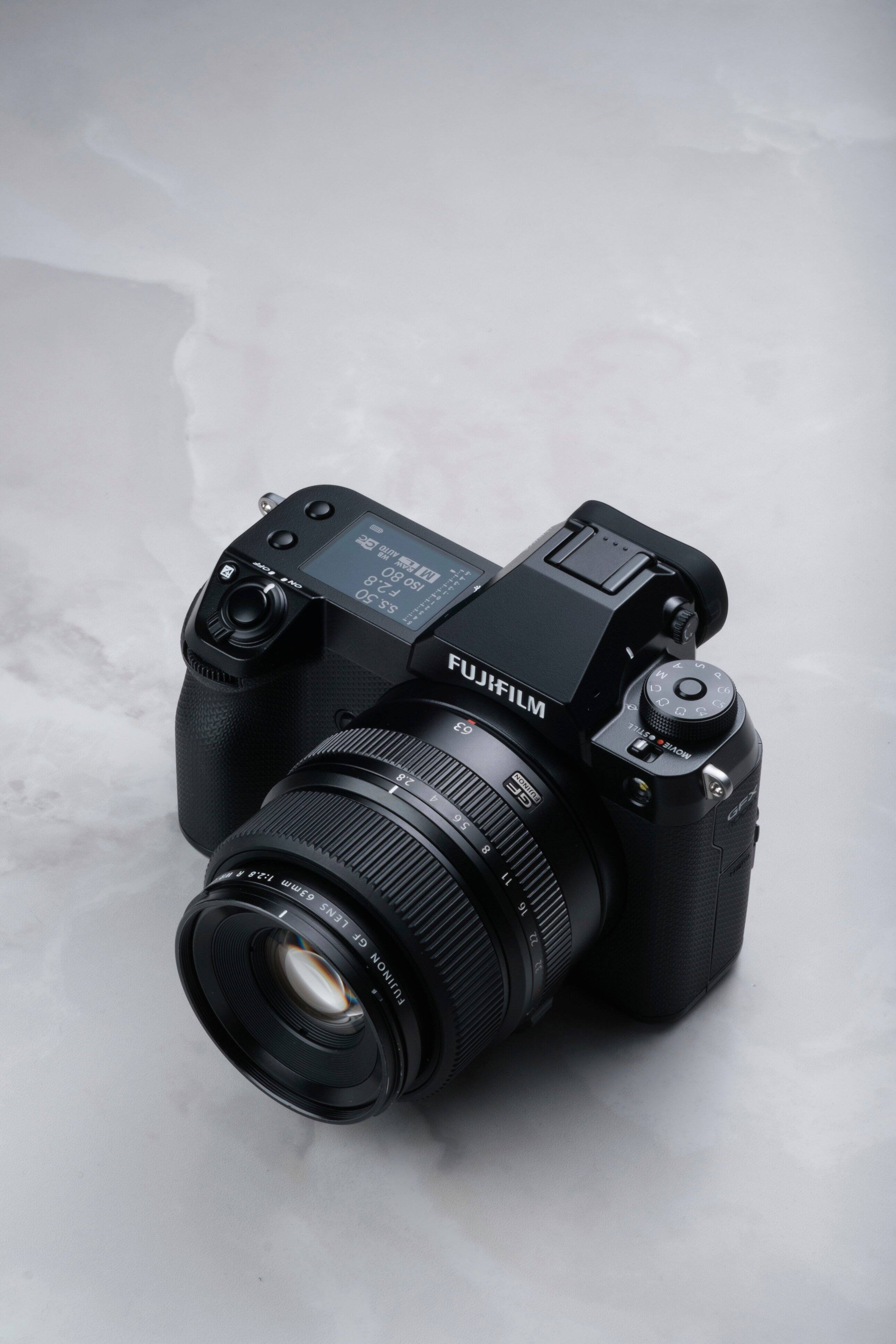 The image features a Fujifilm digital camera with a zoom lens. The camera is positioned on a white surface, possibly a table. The zoom lens is prominently displayed, showcasing its capabilities. The camera is likely used for capturing high-quality images and videos, making it a popular choice for photographers and videographers.