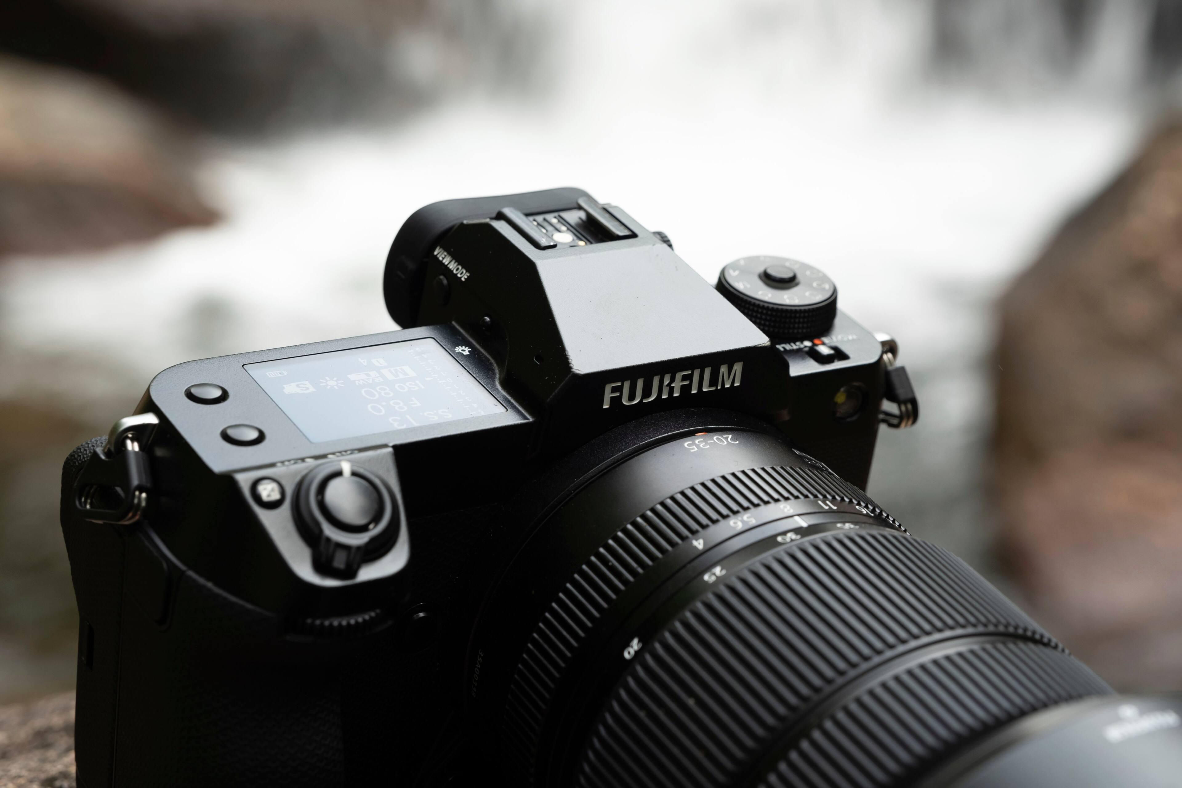 The image features a close-up of a Fujifilm camera, with the lens prominently visible. The camera is positioned in front of a waterfall, creating a captivating scene. The focus of the image is on the camera and its lens, showcasing its features and capabilities.