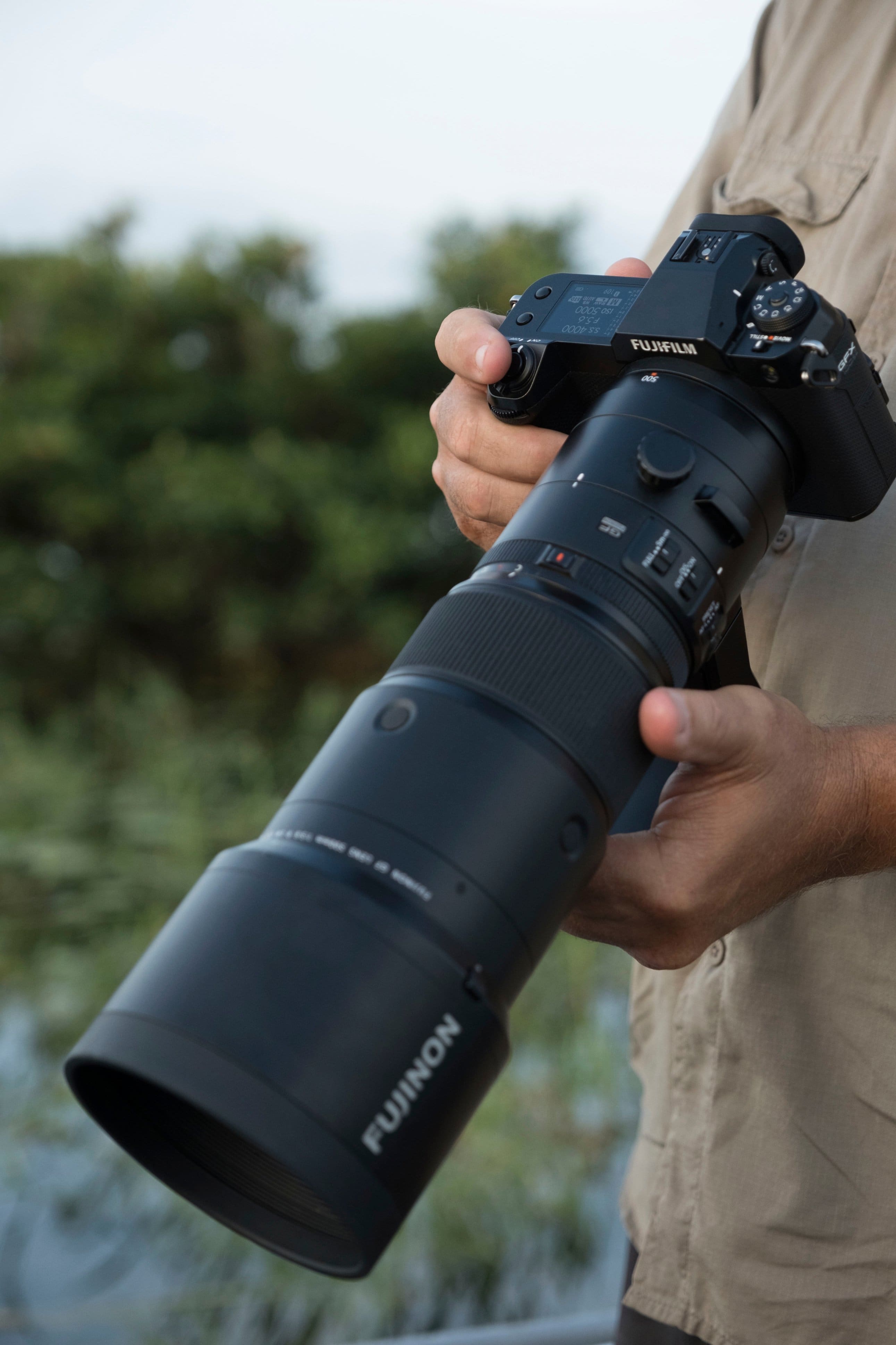 The image features a person holding a large camera, which is a Fujinon lens. The lens is attached to a camera, and the person is likely preparing to take a photograph. The lens is quite large, which suggests that it is designed for capturing high-quality images, possibly in low light conditions or for professional photography.