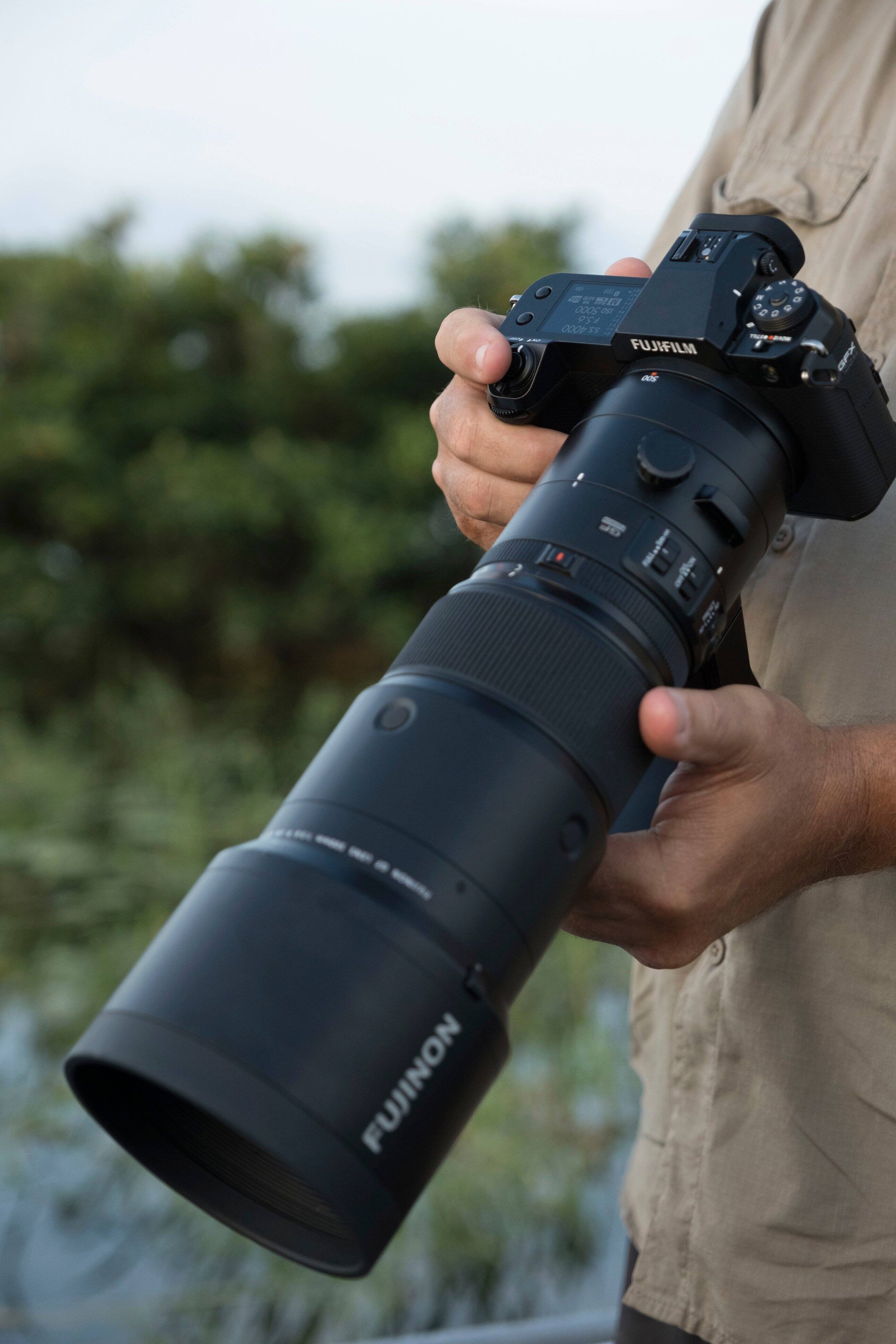 The image features a person holding a large camera, which is a Fujinon lens. The lens is attached to a camera, and the person is likely preparing to take a photograph. The lens is quite large, which suggests that it is designed for capturing high-quality images, possibly in low light conditions or for professional photography.