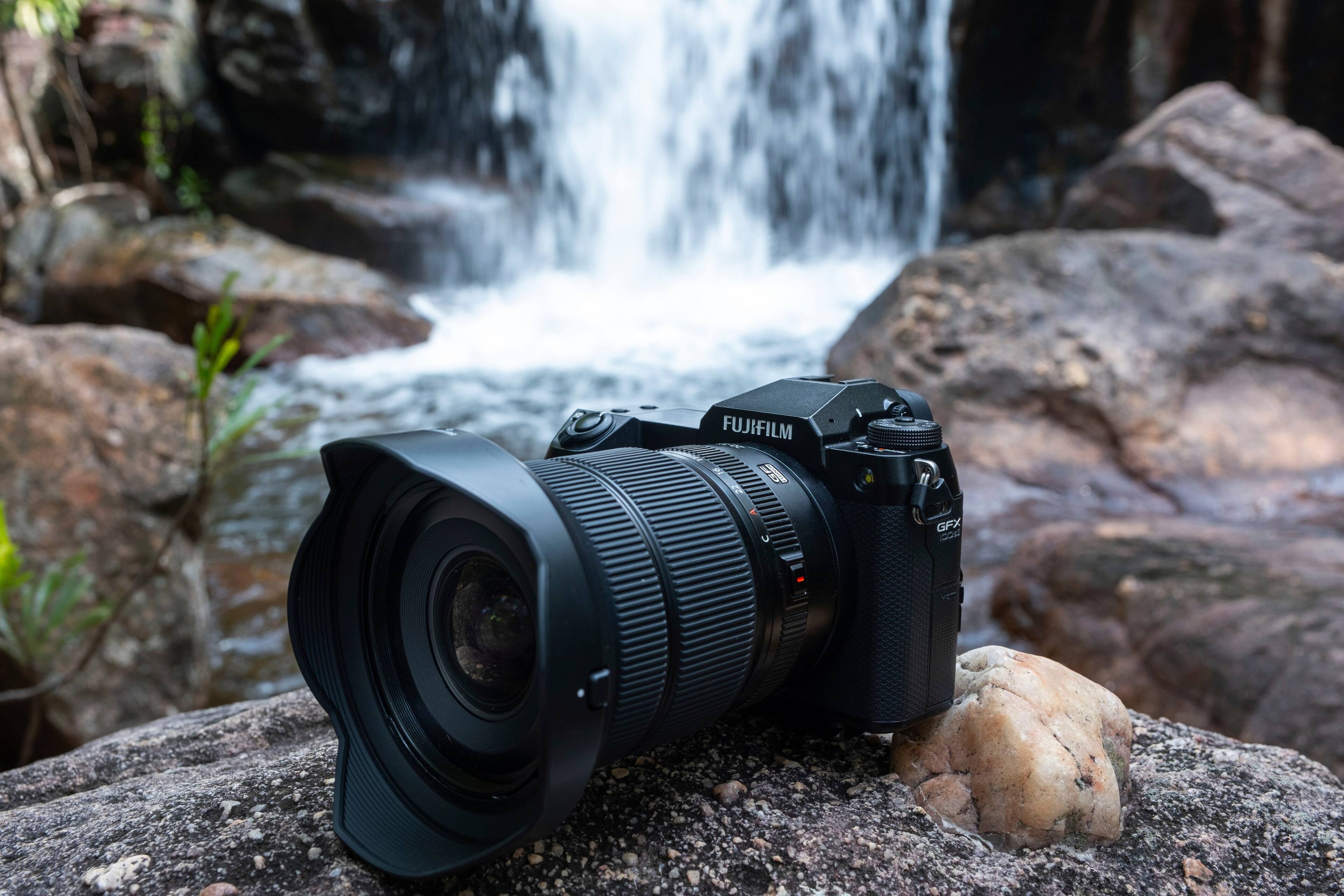 The image features a FUJIFILM E If GFX 100S digital camera, which is positioned on a rock near a waterfall. The camera is facing the water, capturing the stunning scenery. The rock on which the camera is placed is located near the edge of the waterfall, providing a unique perspective of the cascading water. The camera's presence in this natural setting suggests that it is being used to capture the beauty of the landscape, showcasing the capabilities of the FUJIFILM E If GFX 100S in capturing high-quality images in challenging environments.