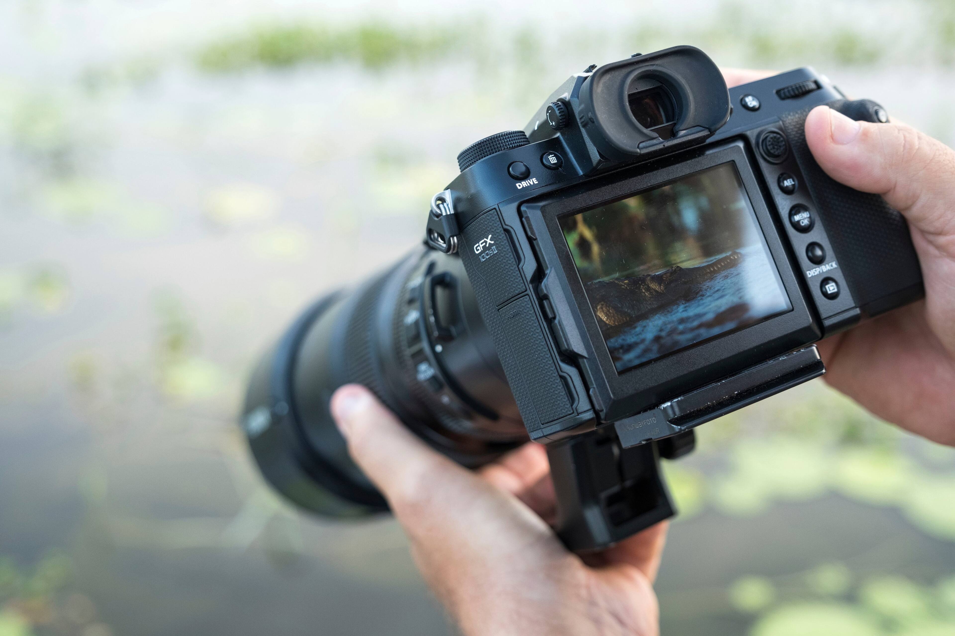 The image features a person holding a digital camera, specifically a Nikon D810, with a lens attached. The camera is being used to take a picture of a pond with lily pads. The person is holding the camera in their hands, focusing on the scene and capturing the moment.