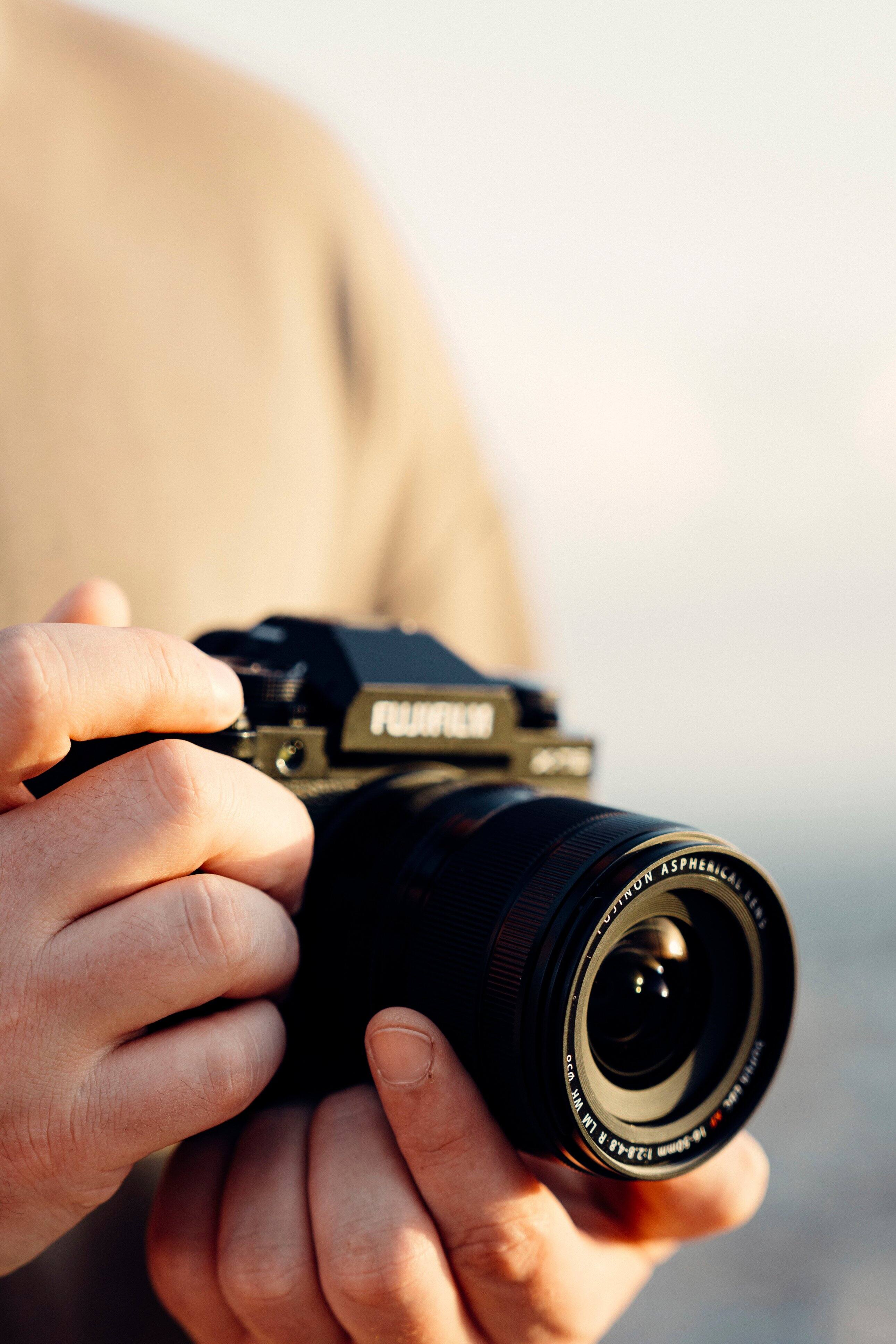 The image features a person holding a Fujifilm camera in their hands. The camera has a large lens, which is a notable feature of this particular model. The person's hands are positioned around the camera, showcasing its size and design. The image is categorized as Digital_Cameras, highlighting the focus on the camera and its features.