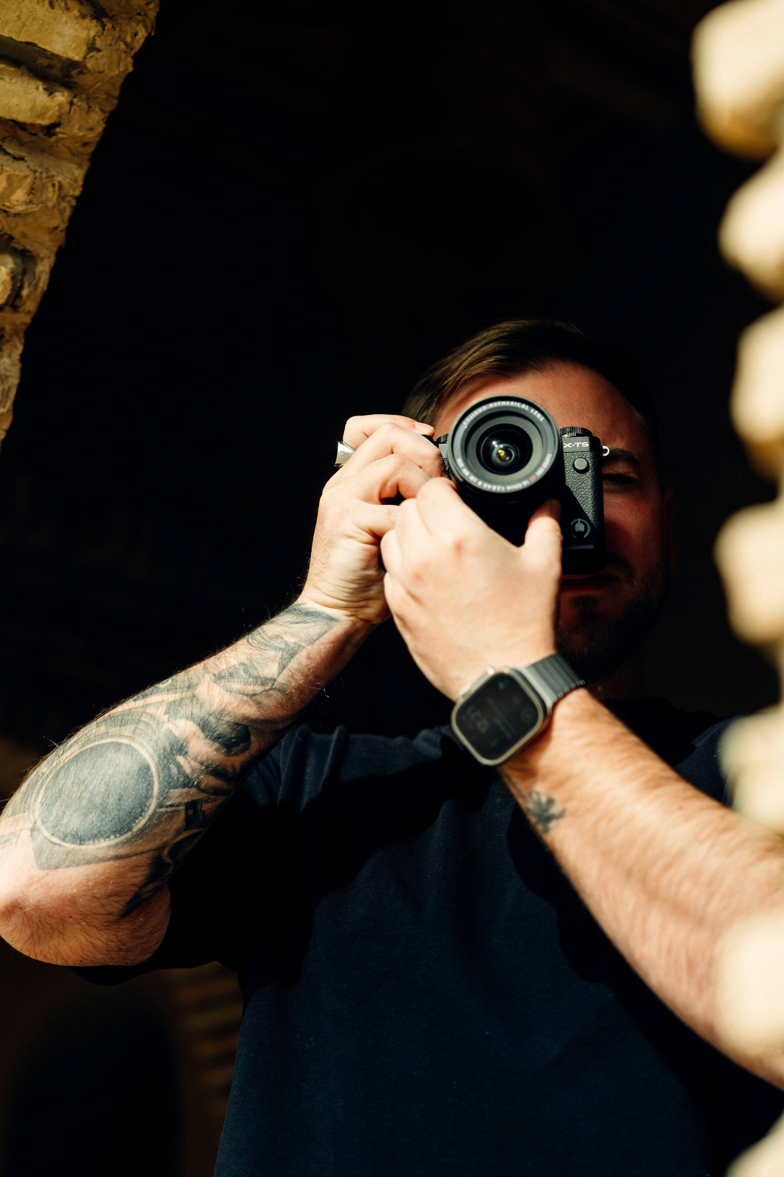 The image features a man holding a camera, likely a digital camera, as he takes a picture. The man is wearing a black shirt and has tattoos on his arms. The camera is positioned in front of him, capturing the moment.