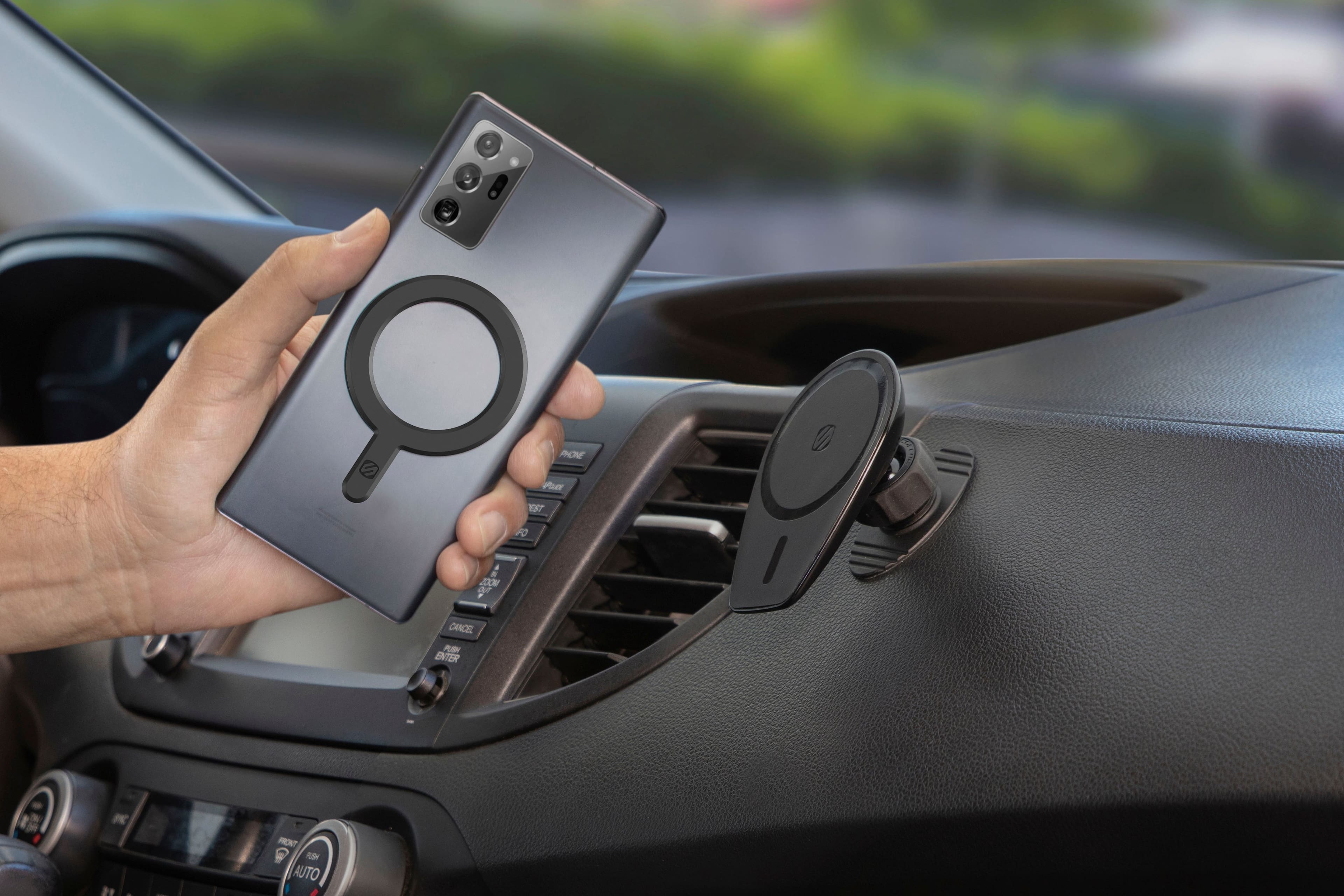 The image shows a person holding a cell phone with a magnifying glass case on it. The magnifying glass case is being used to view the dashboard of a car. The person is holding the cell phone up to the car's dashboard, possibly to take a picture or examine the car's features. The car is parked, and the person is standing near it while using the magnifying glass case on their phone.