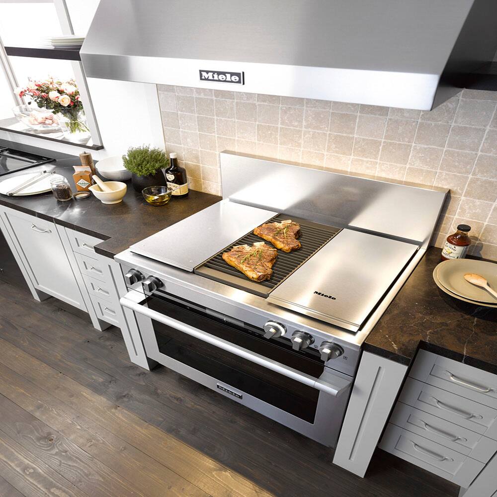 The image shows a kitchen with a stainless steel stove top oven. The oven is currently in use, with two pieces of meat cooking on the grill. The kitchen also features a sink, a counter, and a dining table. Various items can be seen on the counter, such as a bowl, a bottle, and a vase. Additionally, there are two potted plants in the kitchen, one on the counter and another on the dining table.