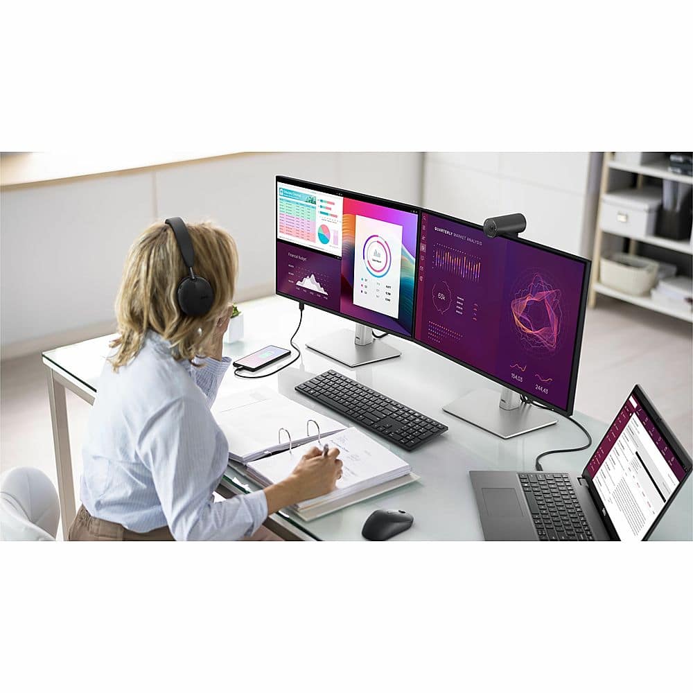 The image features a woman sitting at a desk with a laptop, a keyboard, and a mouse. She is wearing headphones and appears to be working or studying. There are two large computer monitors on the desk, one on the left and one on the right. The woman is also holding a book, which is placed on the desk near her. The scene suggests a productive work environment, possibly in an office or study area.