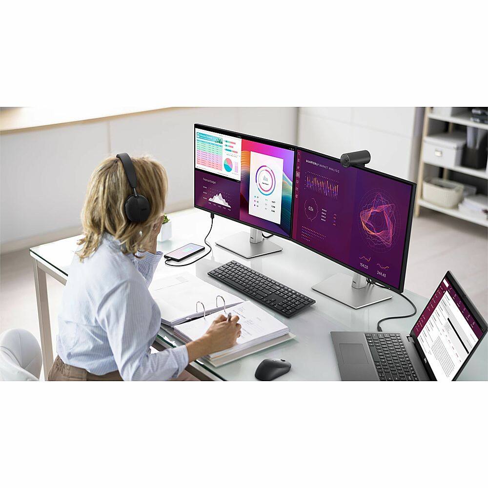 The image features a woman sitting at a desk with a laptop, a keyboard, and a mouse. She is wearing headphones and appears to be working or studying. There are two large computer monitors on the desk, one on the left and one on the right. The woman is also holding a book, which is placed on the desk near her. The scene suggests a productive work environment, possibly in an office or study area.