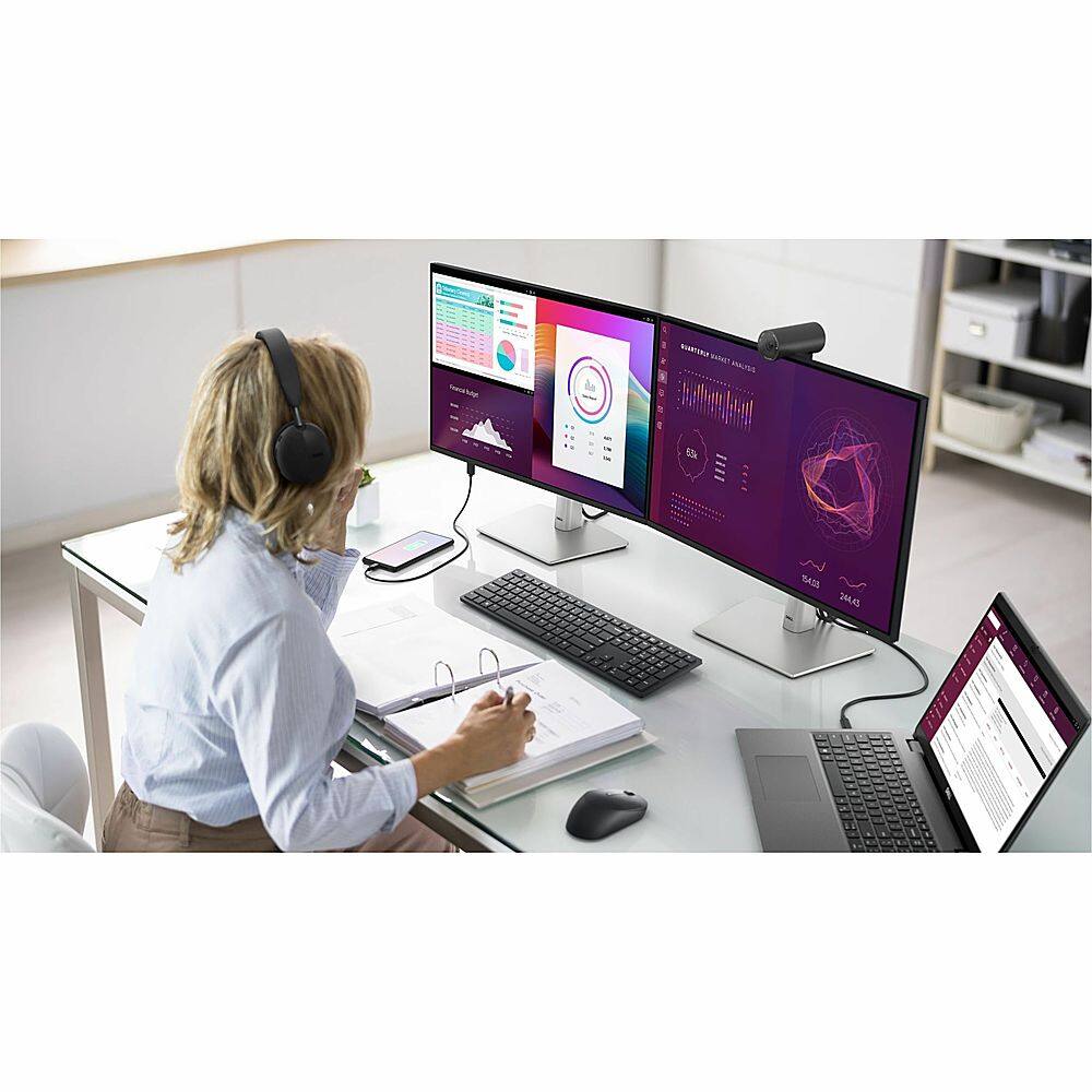 The image shows a woman sitting at a desk with a laptop, a keyboard, and a mouse. She is wearing headphones and appears to be working or studying. There are also two books on the desk, one of which is a spiral-bound book. The woman is surrounded by a comfortable environment, with a couch and a chair nearby.