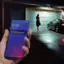 The image shows a person holding a cell phone with the time displayed as 12:45. The phone is being used to view a message that says "NIGHT OWL Motion Has Been Detected (Home) Floodlight Camera Saw Something." The scene takes place in front of a garage, with a car parked nearby.