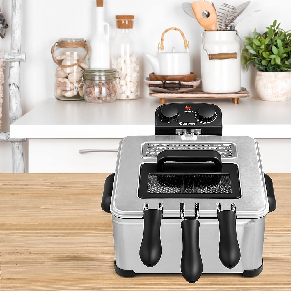 The image features a kitchen counter with a silver toaster oven sitting on top of the counter. The toaster oven has a black handle and is placed next to a cutting board. There are various kitchen items around the toaster oven, including a bottle, a cup, a knife, and a spoon. Additionally, there are two potted plants in the scene, one on the left side and another on the right side of the counter.