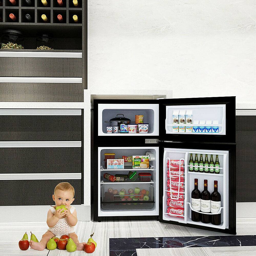 The image shows a baby sitting on the floor in front of an open refrigerator. The refrigerator is filled with various food items, including apples and oranges. There are also several bottles of wine placed in the refrigerator. The baby is holding a piece of fruit, possibly an apple, while sitting on the floor. The scene appears to be set in a kitchen, with the refrigerator being the main focus of the image.