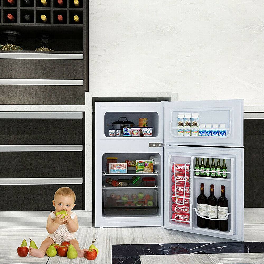 The image features a baby sitting on the floor in front of an open refrigerator. The refrigerator is filled with various food items, including several bottles of wine and a few apples. The baby appears to be playing with the apples, and there is also a bowl placed nearby. The scene creates a playful and whimsical atmosphere, as the baby interacts with the refrigerator and its contents.