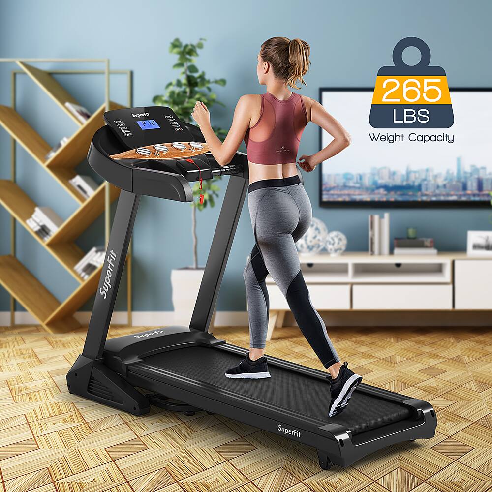 The image features a woman exercising on a treadmill in her living room. She is wearing a tank top and leggings, and the treadmill is set at a 12% incline. The room has a TV on the wall, and there are several books scattered around the area. The treadmill is designed to hold up to 265 lbs, making it suitable for a wide range of users.