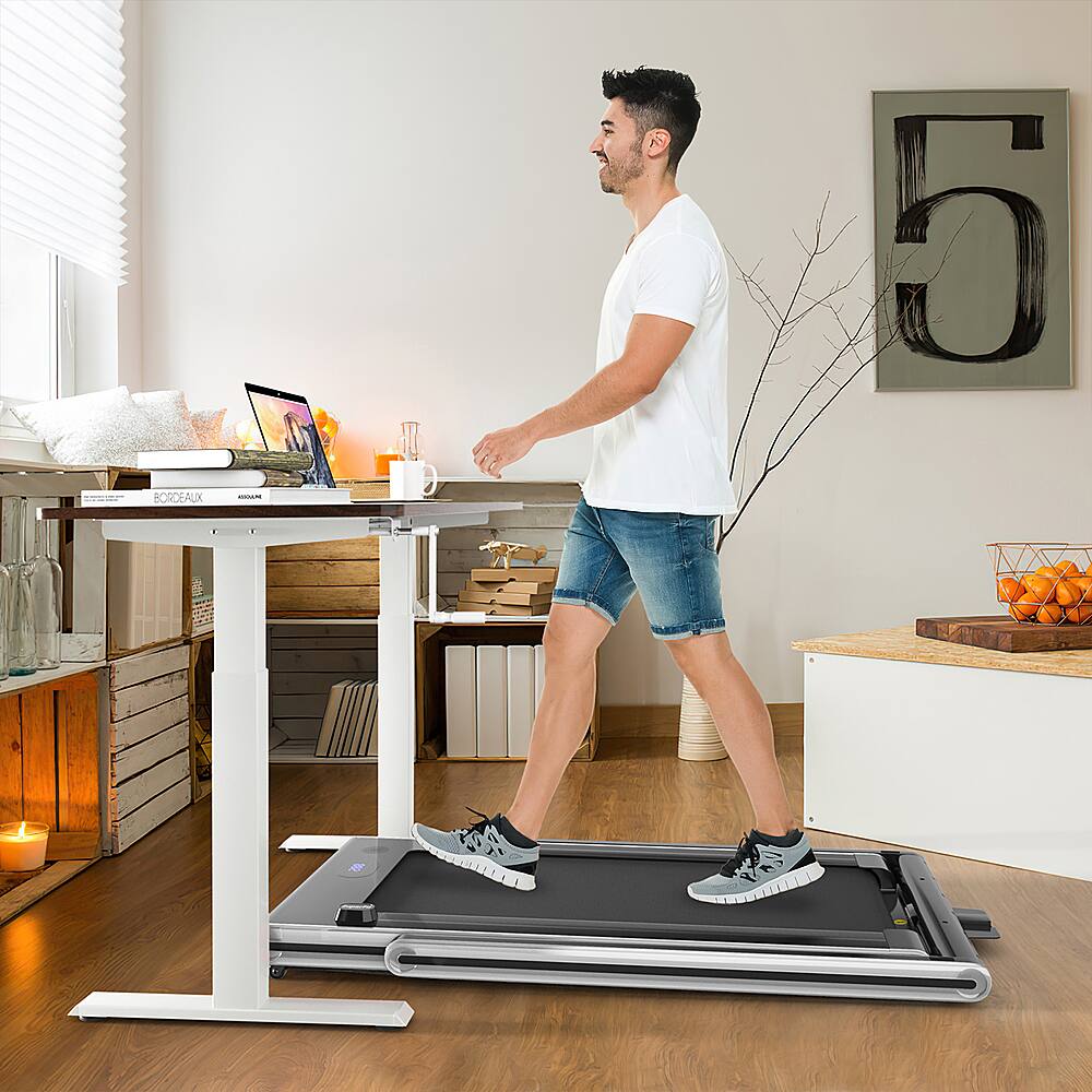 The image shows a man standing on a treadmill in a living room. He is wearing a white shirt and blue shorts, and he is using a laptop while standing on the treadmill. The room also contains a dining table, a few books, and a vase. There are also some oranges in the room, adding a touch of color to the scene.