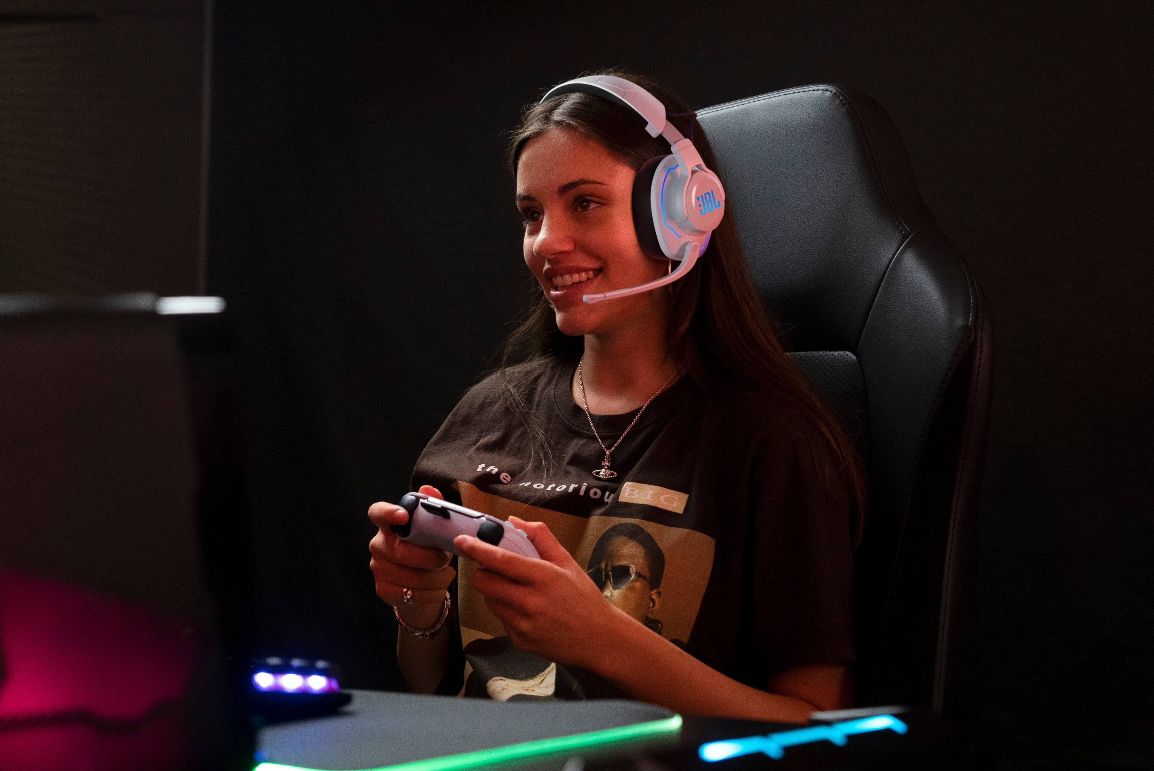 The image shows a woman sitting in a chair, wearing headphones and holding a game controller. She is smiling and appears to be enjoying her gaming session. The headphones are positioned on her head, and the controller is held in her hand. The woman is the main focus of the image, and her engagement in the gaming activity is evident.