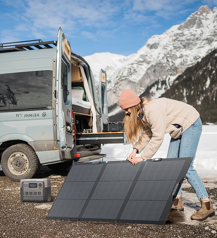 I cannot group or correct the text as it appears to be unrelated to the image. The image shows a woman standing next to a solar panel, possibly preparing to set up camp. The van is parked nearby, and there is a radio on the ground. The woman is wearing a beanie and a jacket, and she is looking at the solar panel.