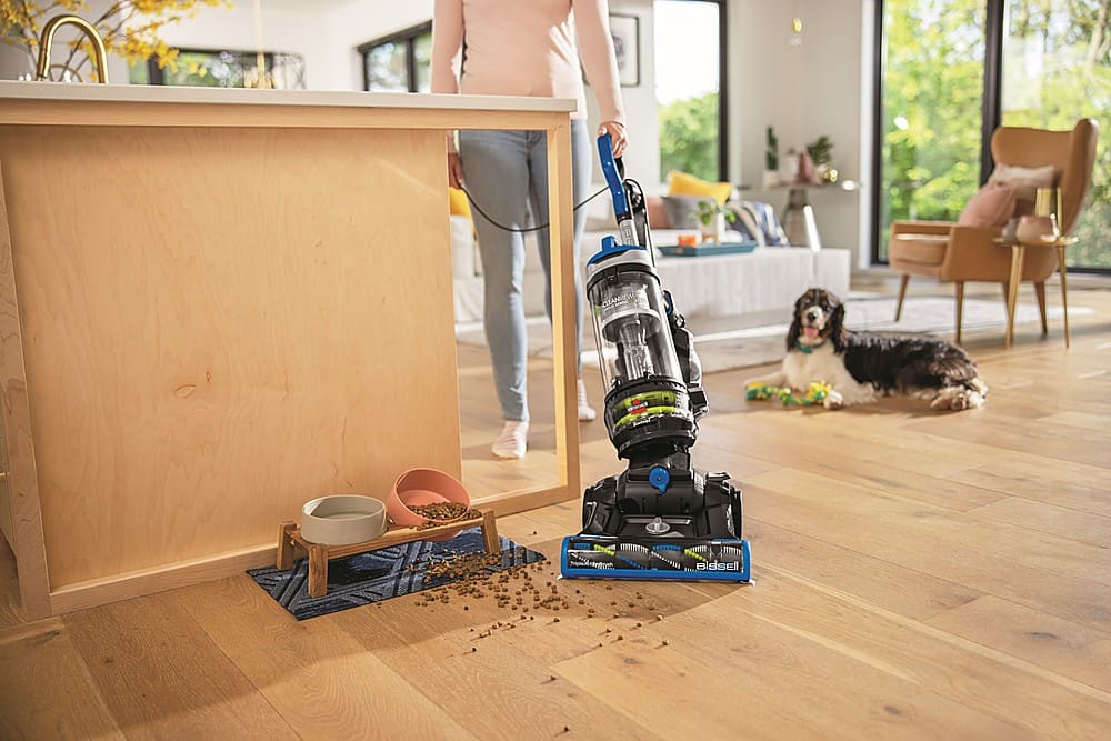 The image shows a woman vacuuming a hardwood floor in a living room. She is using a robotic vacuum cleaner, which is positioned near the center of the room. The room is furnished with a couch, a chair, and a dining table. There is also a dog lying on the floor, possibly watching the woman as she cleans the area.