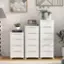 The image features a white dresser with three drawers, placed in a corner of a room. On top of the dresser, there is a radio, a vase with a plant, and a small doll. Additionally, there are two books placed on the dresser, along with a potted plant nearby. The overall scene appears to be a cozy and organized living space.