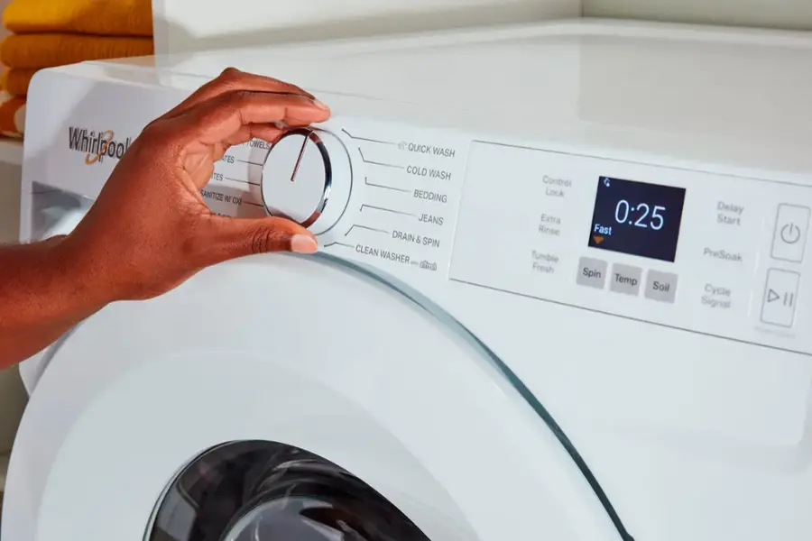 While looking at the washer, the person is pressing the "Quick Wash" button. The washer has various options, including "Cold Wash," "Bedding," "Jeans," "Drain & Spin," "Clean Washer," and "Extra Fast 0:25." Other buttons include "Tumble Fresh Spin," "Temp Soil," "Delay Start," "PreSoak Cycle," and "Signal Hold to Start."