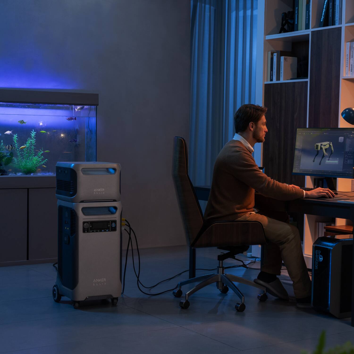 The man is sitting at a desk, using a computer. He is wearing a brown sweater and is positioned in front of a monitor. There is a fish tank nearby, and a bookshelf with books in the background. A keyboard and mouse are also present on the desk.