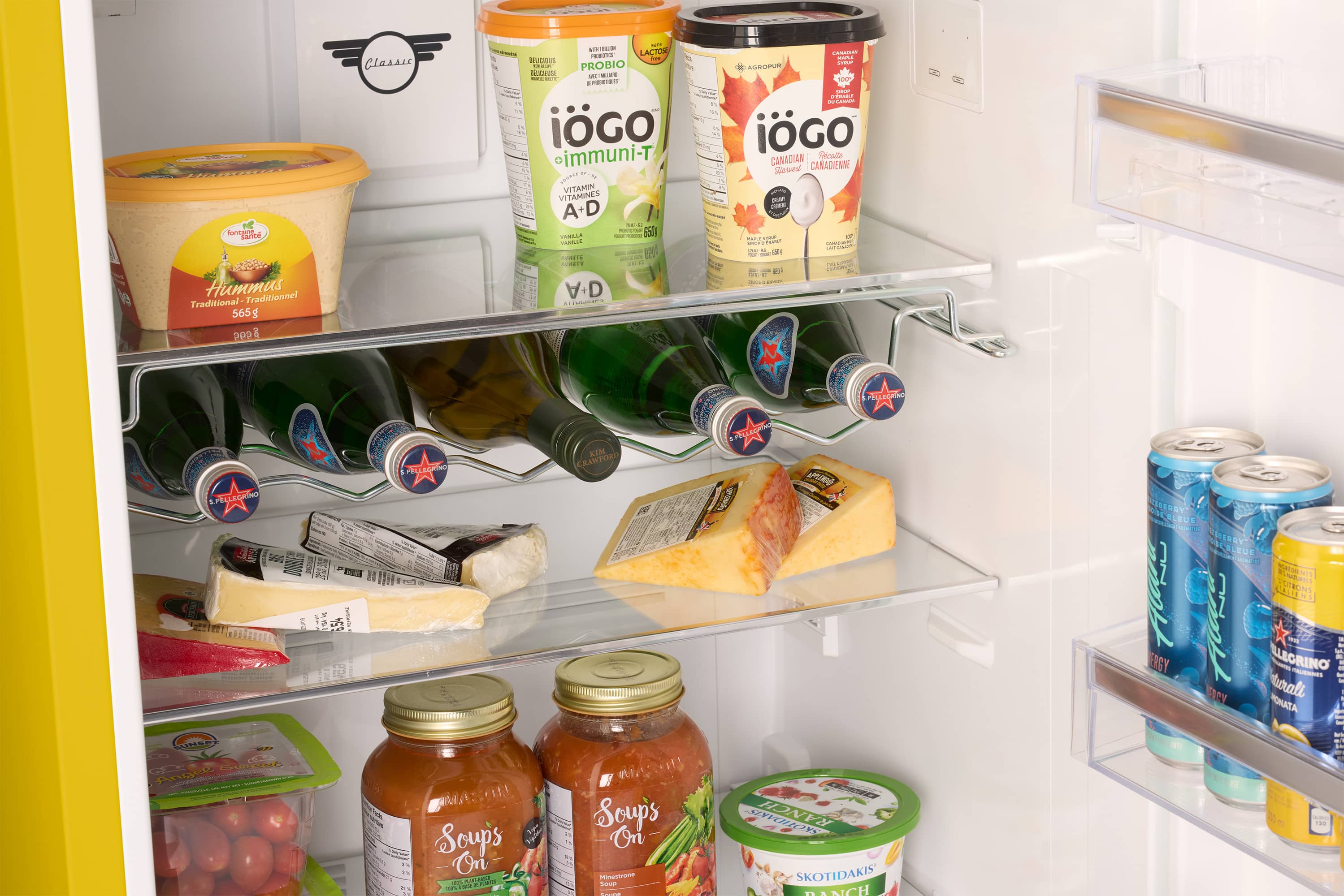 The image shows a refrigerator with various food items and drinks. The refrigerator contains a bottle of hummus, a bottle of yogurt, and a bottle of juice. There are also multiple cans of soda, including some on the top shelf and others on the bottom shelf. Additionally, there are two bowls inside the refrigerator, one placed on the top shelf and the other on the bottom shelf. The refrigerator is well-stocked with a variety of food and drinks to choose from.