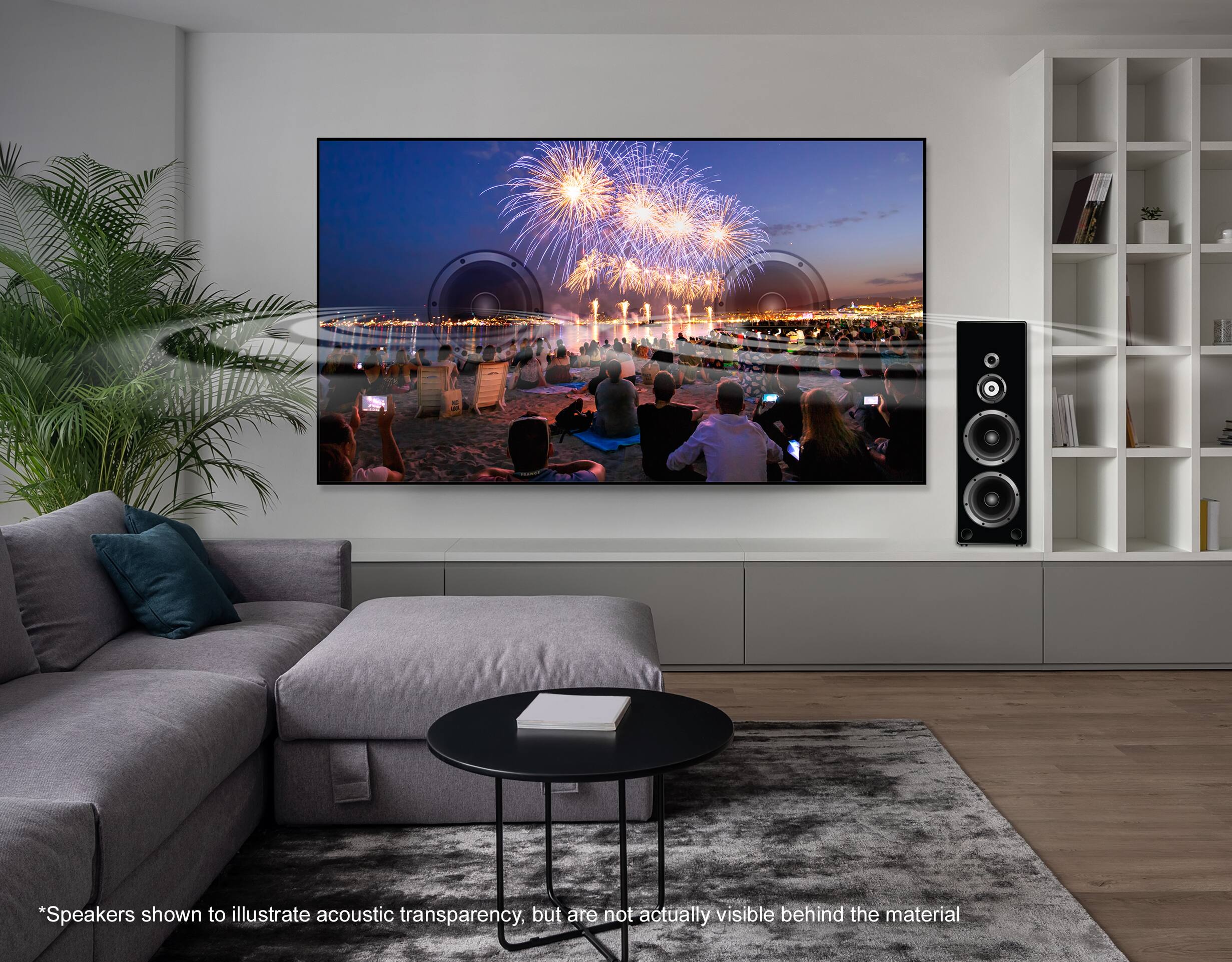The image features a large flat screen TV mounted on a wall, displaying a fireworks show. The room is furnished with a couch and a coffee table, and there are several books and potted plants in the space. The TV is described as having speakers, but they are not actually visible behind the material. The image is meant to showcase the acoustic transparency of the material, but the speakers are not physically present.