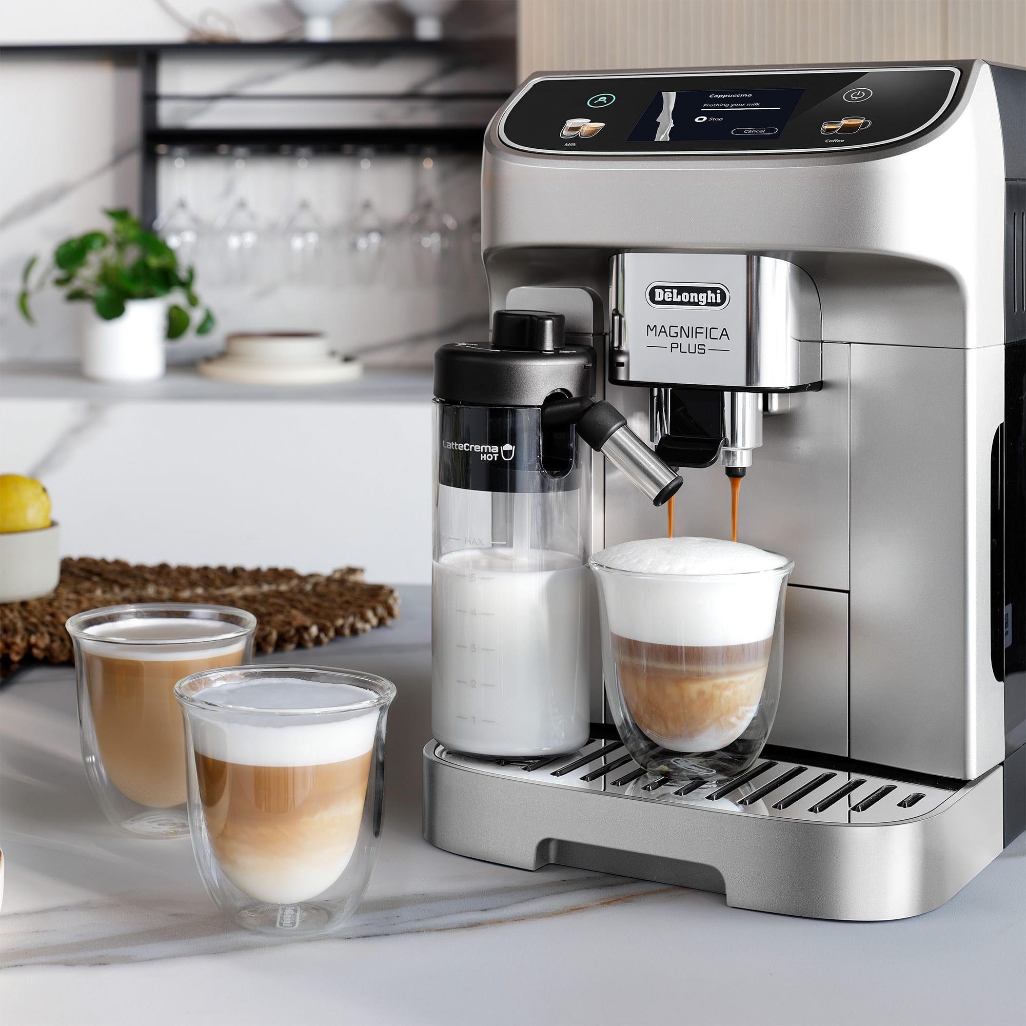 The image shows a Longhi Magnifica Plus espresso machine with a cup of coffee being poured into a glass. The machine is located on a counter with a few cups and wine glasses nearby. The coffee is being made with a creamer, and there are also some oranges present in the scene. The espresso machine is a modern and sleek appliance, and it is likely used to make a variety of coffee drinks, such as cappuccinos and lattes.