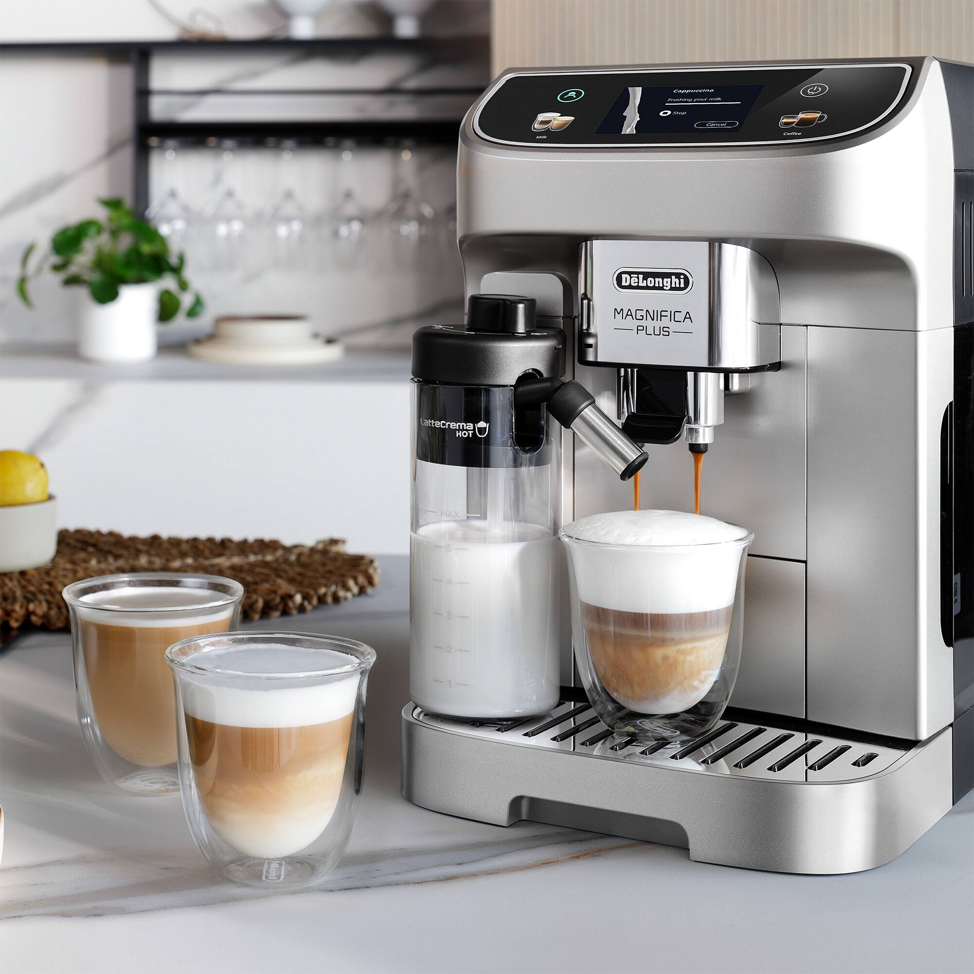 The image shows a Longhi Magnifica Plus espresso machine with a cup of coffee being poured into a glass. The machine is located on a counter with a few cups and wine glasses nearby. The coffee is being made with a creamer, and there are also some oranges present in the scene. The espresso machine is a modern and sleek appliance, and it is likely used to make a variety of coffee drinks, such as cappuccinos and lattes.