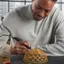 "A man is smiling and holding a small yellow object in his hands. He is using a tool to work on the object, possibly crafting or fixing it. The man is wearing a gray shirt and appears to be enjoying the process."