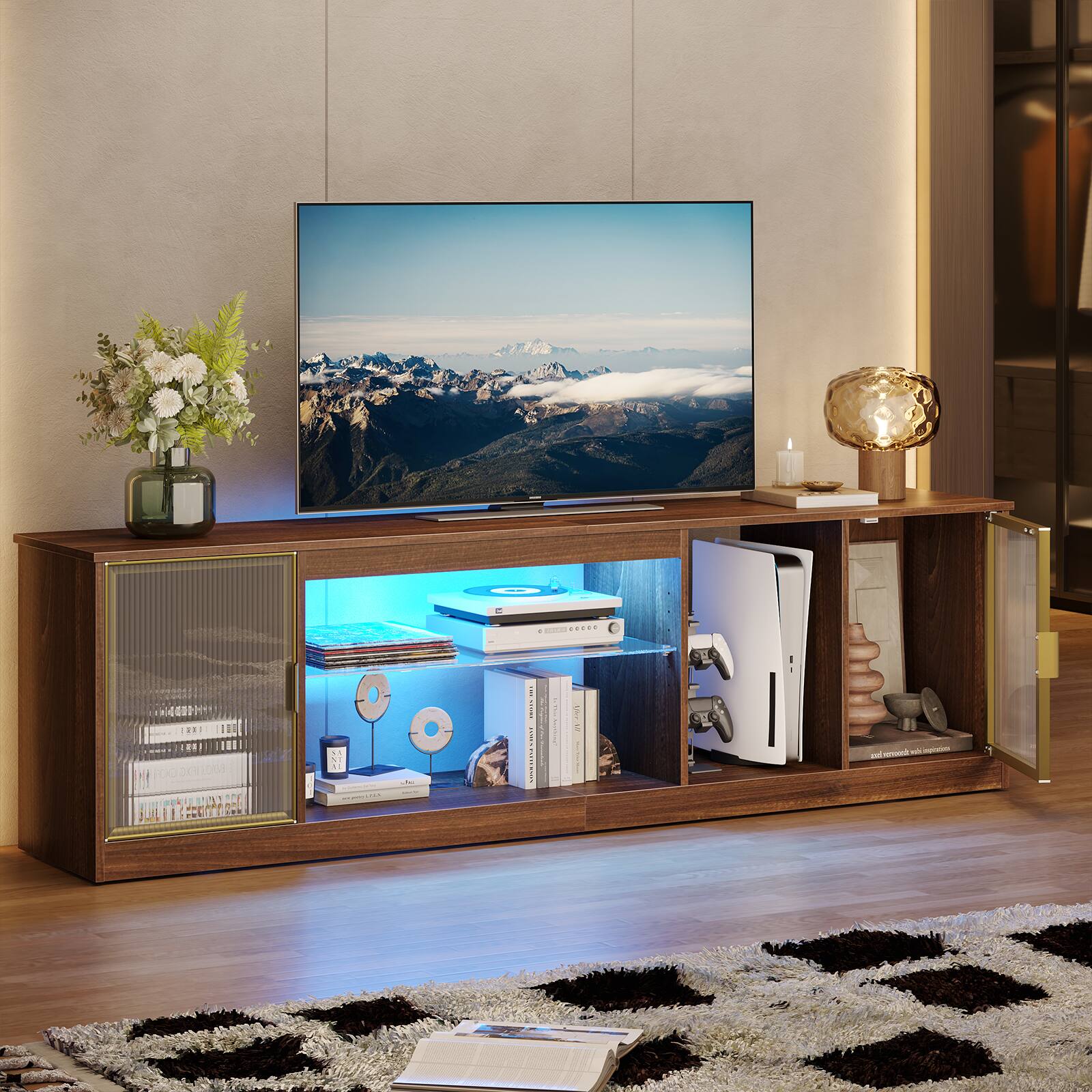 The image features a living room with a flat screen TV on top of a wooden stand. The TV is turned on, displaying a beautiful mountain scene. The room also contains a wooden entertainment center with a variety of items, including a vase, a potted plant, and several books. There are also two remotes placed nearby, likely for controlling the TV and other electronic devices. The overall setting appears to be a cozy and inviting space for relaxation and entertainment.