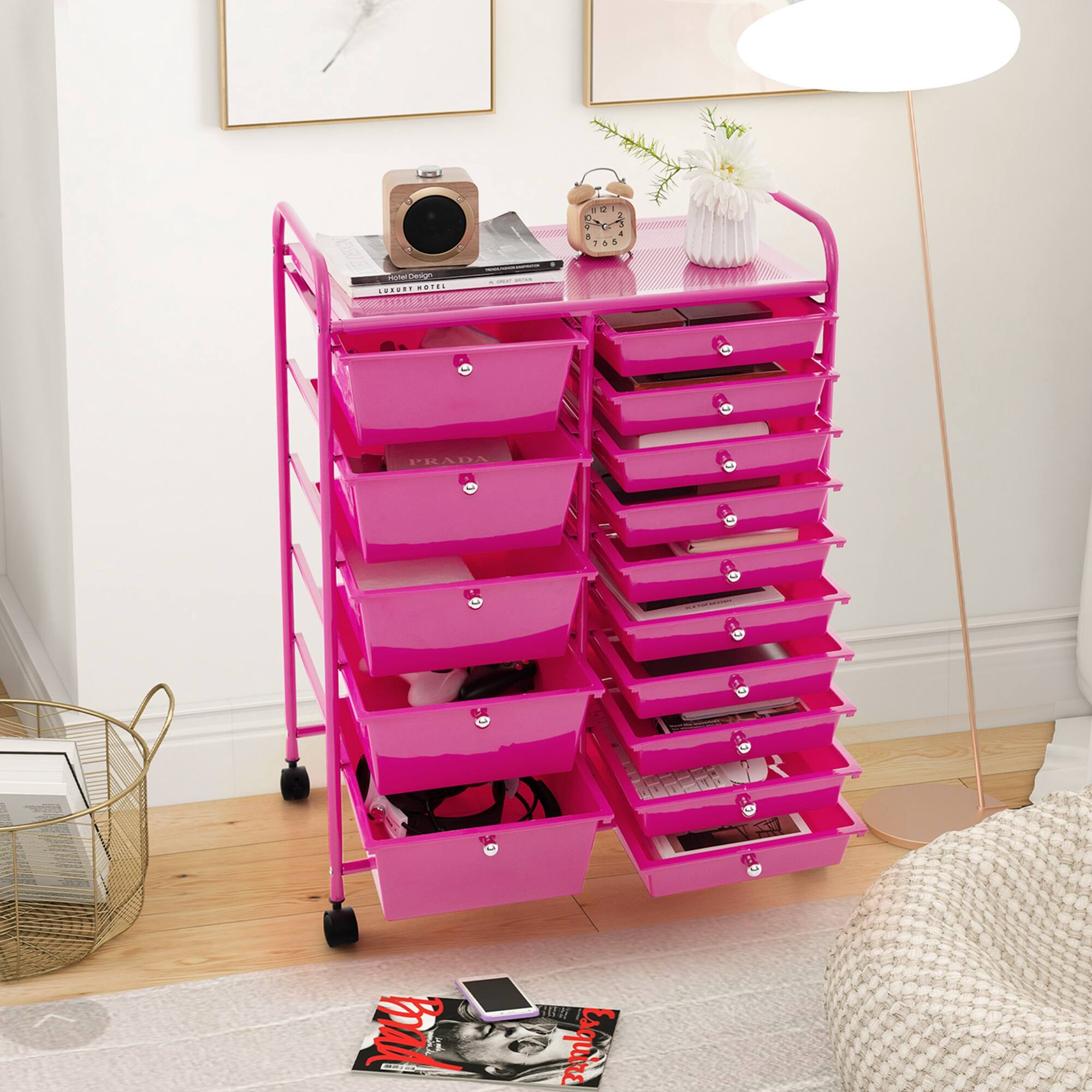 The image features a pink cart with a variety of items on it. There are several books, a cell phone, and a vase placed on the cart. The cart is located in a room with a wooden floor, and there is a potted plant nearby. The scene also includes a bed, a chair, and a couch. Additionally, there are two cups and a remote control in the room.