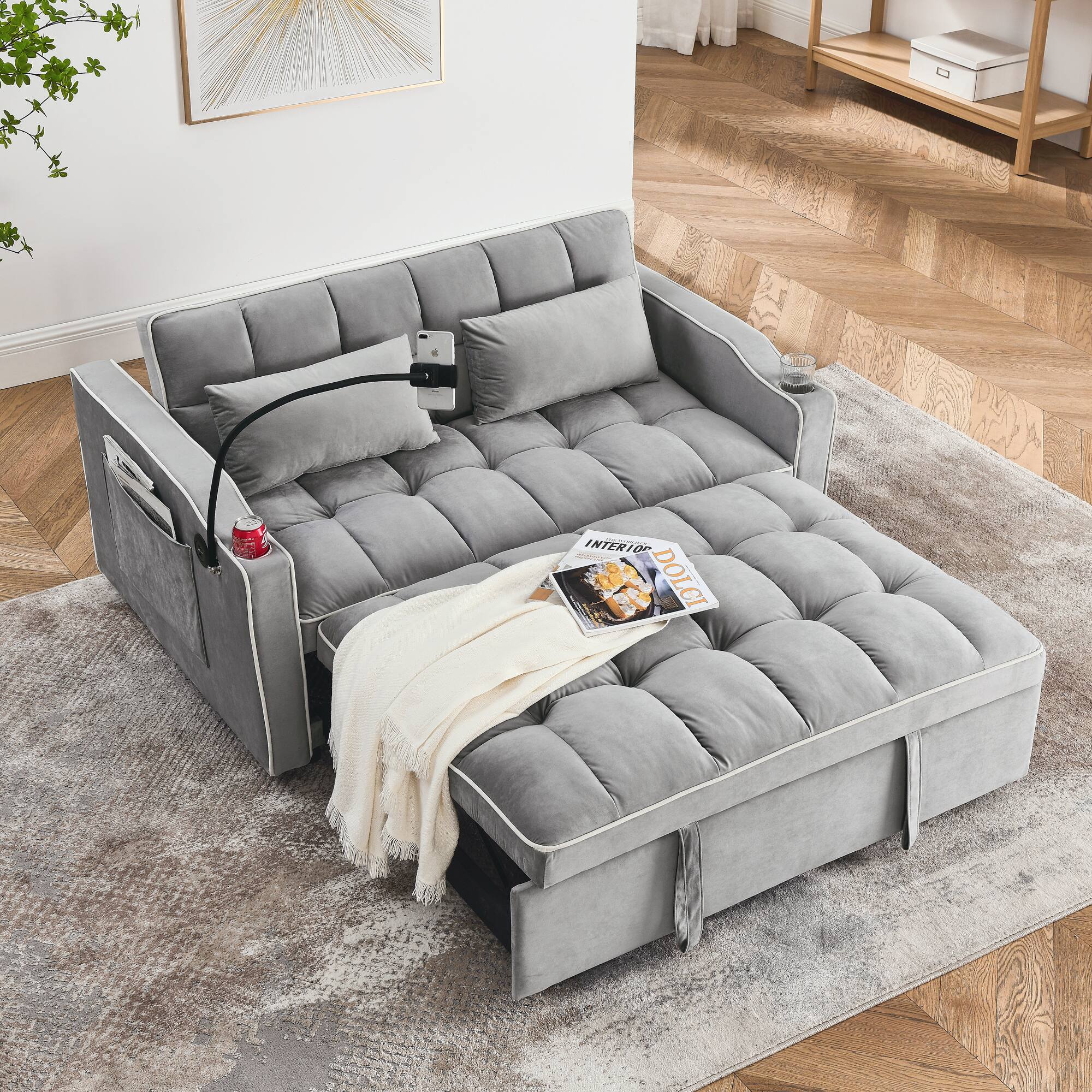 The image shows a modern, gray upholstered sofa with a tufted design, placed on a textured rug in a room with wooden herringbone flooring. The sofa has a sleek, contemporary look with clean lines and a minimalist aesthetic. A white throw blanket is casually draped over one side, and a magazine with the visible text "INTERIOR DOLCI" is placed on the seat. A smartphone is mounted on a flexible stand attached to the sofa, and a red can is on the armrest. The background features a framed abstract artwork on the wall and a wooden side table with a small box on it.