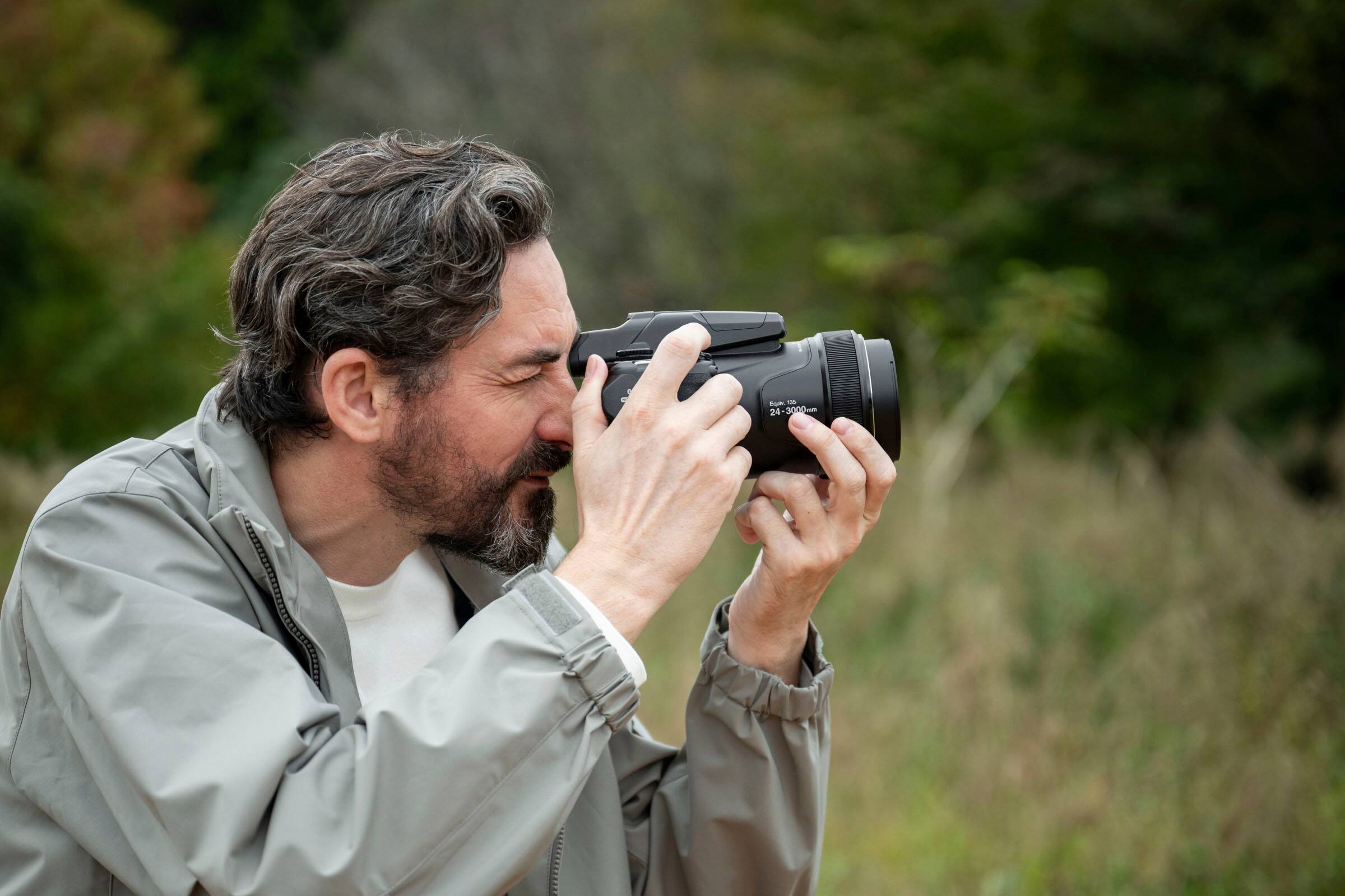 The man is holding a camera that has a zoom lens with a range of 24-3000mm.